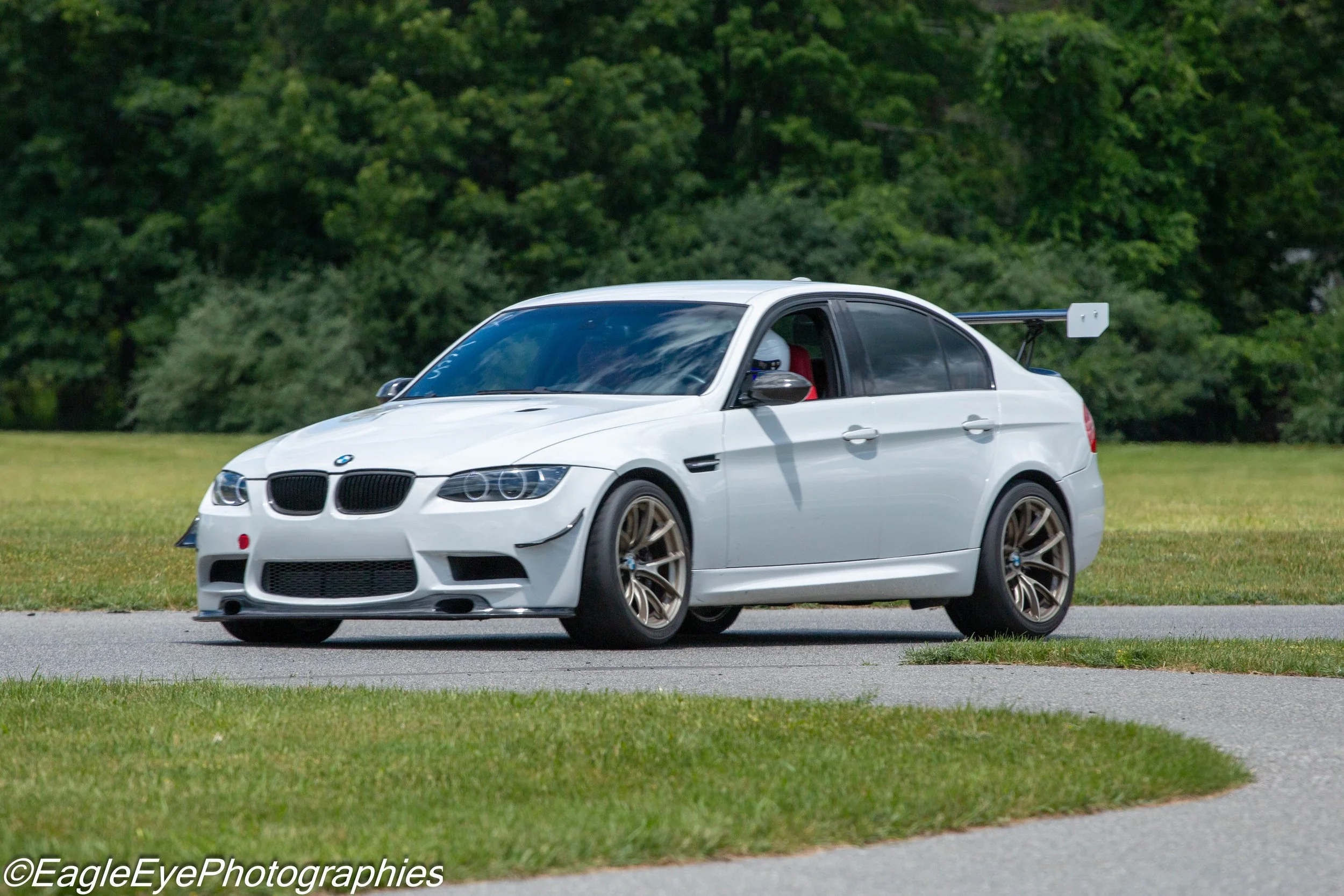 A white BMW sports car with a large rear wing is parked on a grassy area, with a background of green trees.