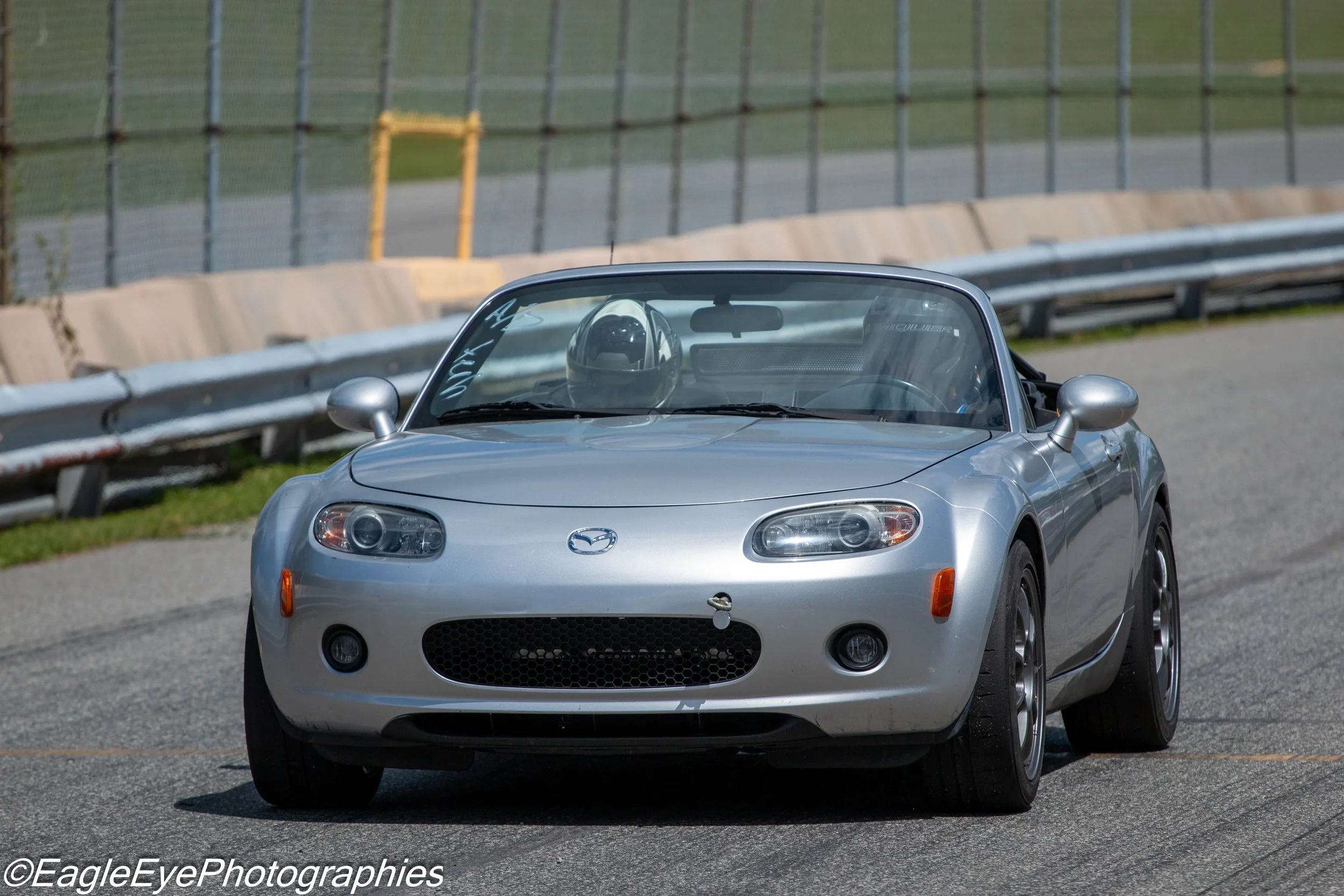 A silver Mazda convertible car on a race track, with a driver wearing a helmet visible through the windshield, surrounded by safety barriers and fencing.