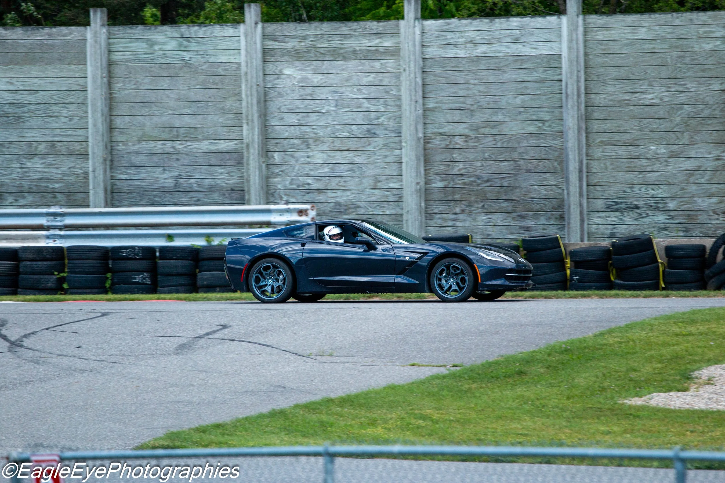 A dark blue sports car with a driver wearing a helmet, driving on a racetrack with safety barriers and a wooden fence in the background.