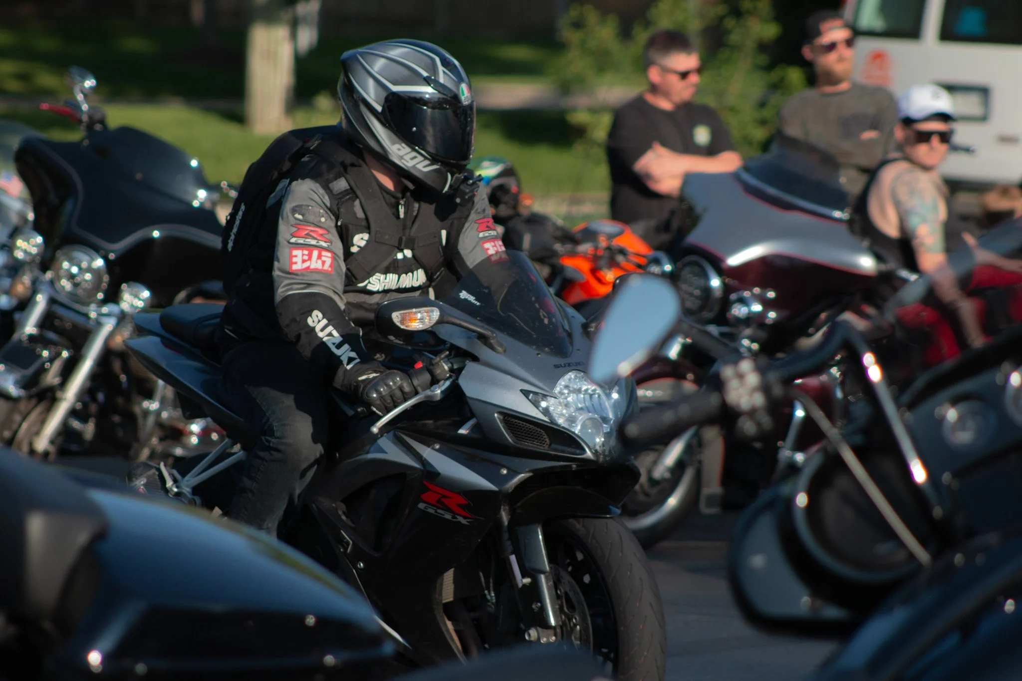 Motorcycle riders parked outdoors, one wearing a black helmet and gear, others in sunglasses and casual attire, with motorcycles lined up.