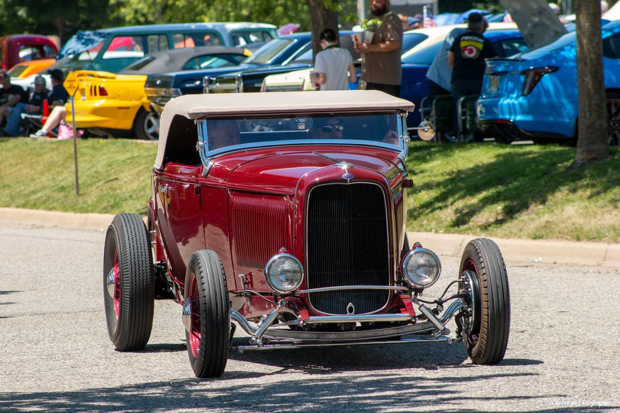 A vintage red car with beige top driving on a street during an outdoor car show, with classic cars and people in the background.