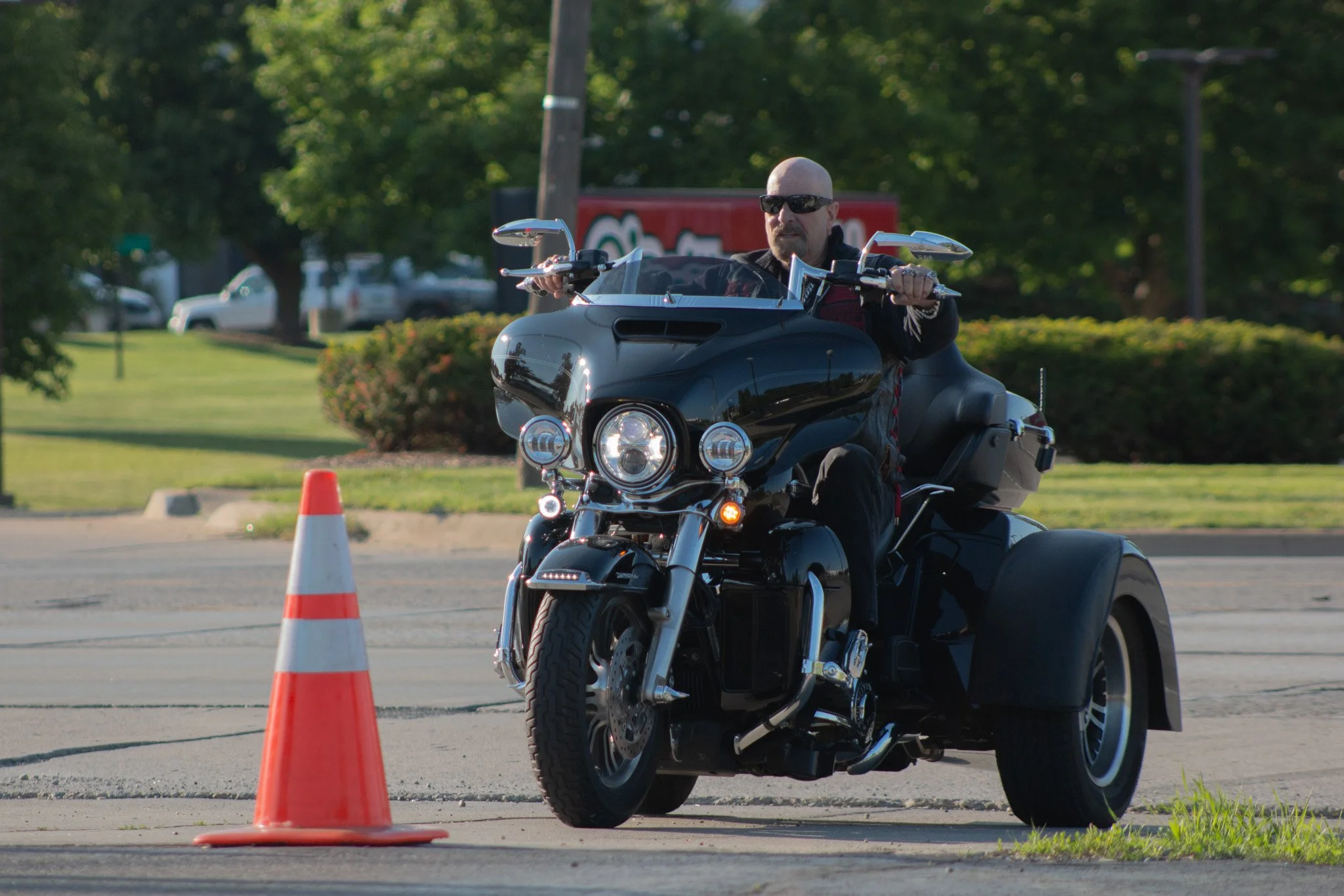 A man riding a black motorcycle navigates through orange traffic cones in an outdoor parking lot, with trees and parked cars in the background.