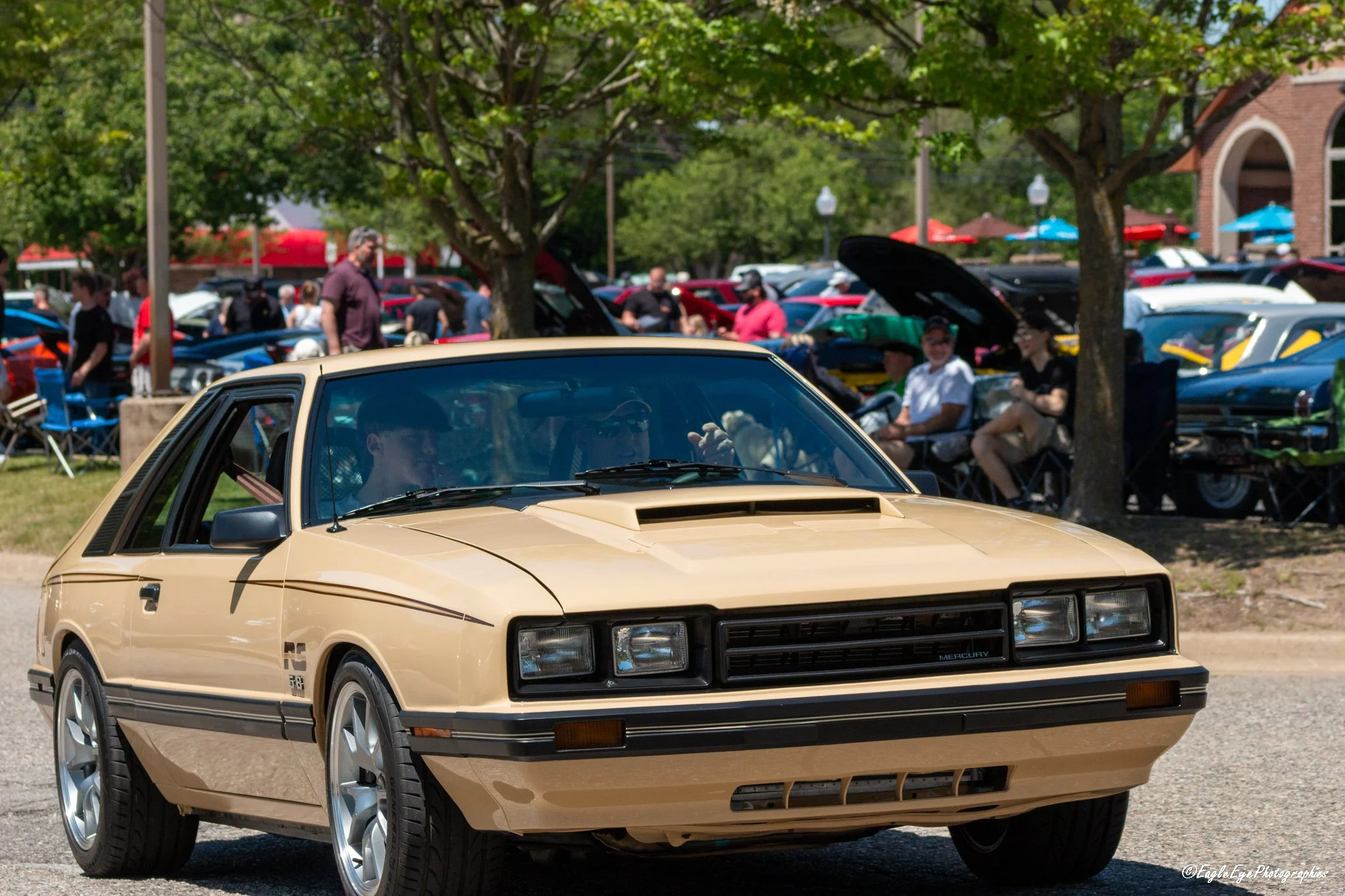 A vintage beige car parked in front of a busy car show or outdoor gathering with people. The background shows trees, multiple cars including some with open hoods, and colorful umbrellas.