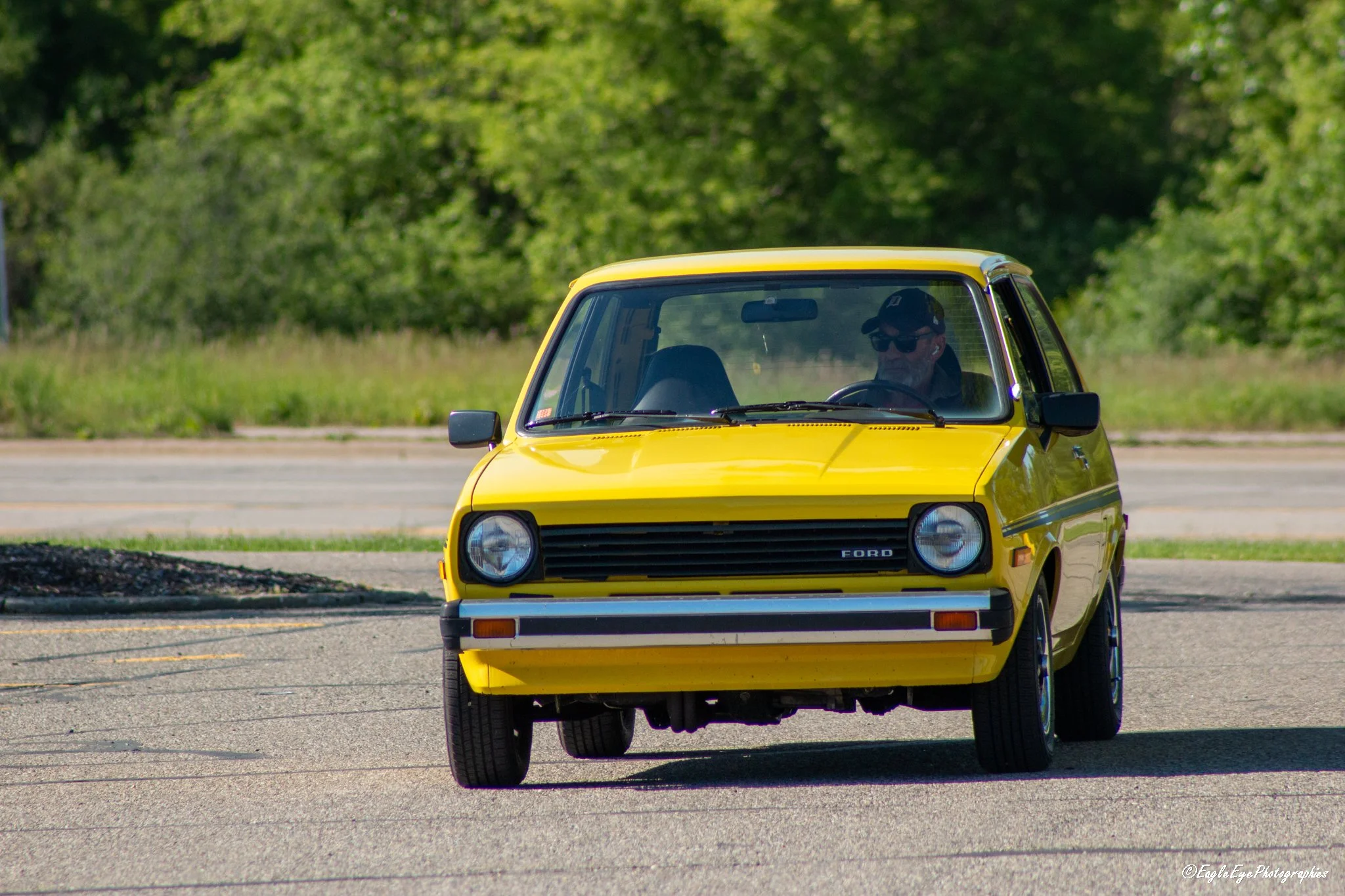 A yellow vintage Ford car parked on a paved surface with a driver wearing sunglasses and a cap inside. Green trees are blurred in the background.