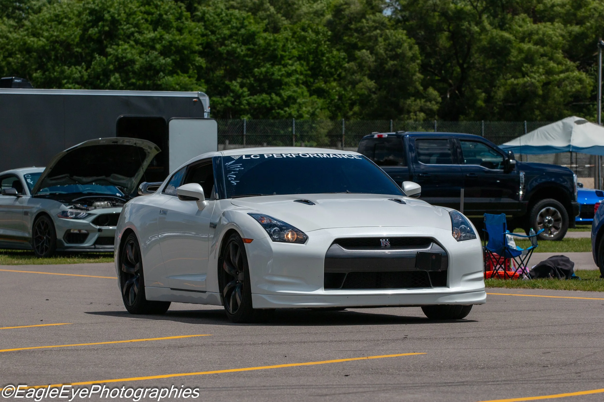 A white Nissan GT-R sports car with a black front grille parked at an outdoor car event. Behind it, a silver car with its hood open, a black pickup truck, a large black trailer, and a blue tent are visible. The background features green trees and a c