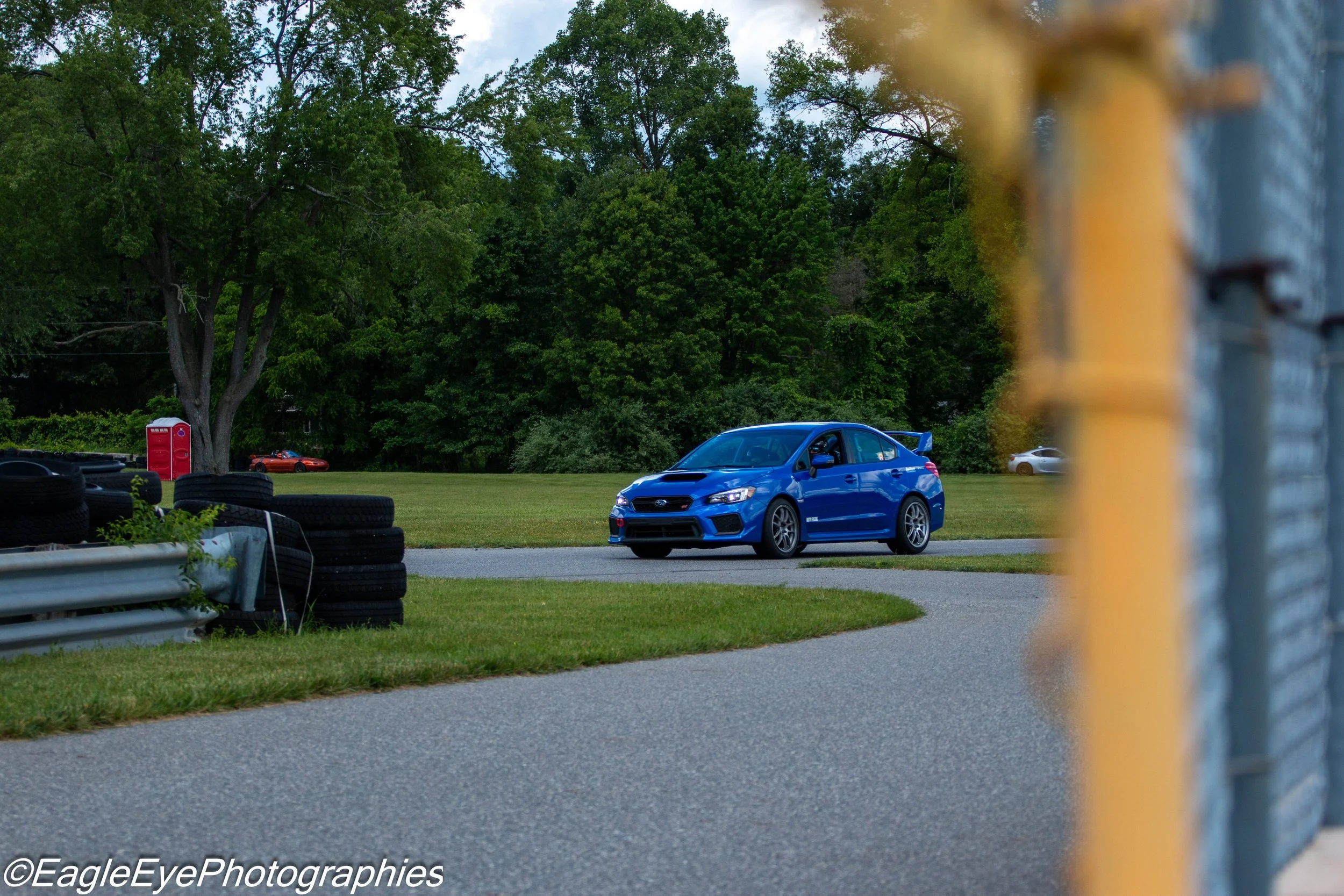 A bright blue Subaru sports car parked on a grassy area near trees with a red phone booth and some tires in the background, viewed through a yellow and gray metal fence.