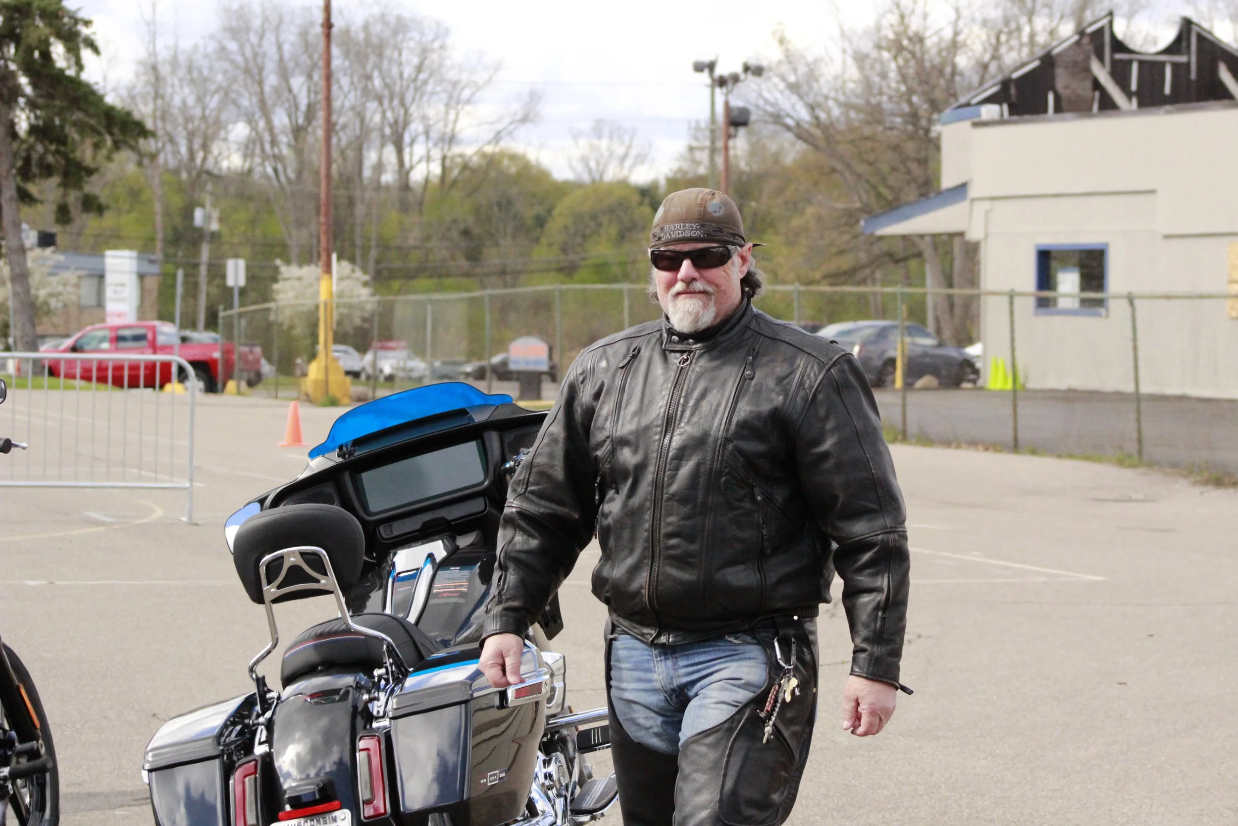 A man in a black leather jacket and sunglasses standing next to a blue and black motorcycle in a parking lot with trees and buildings in the background.