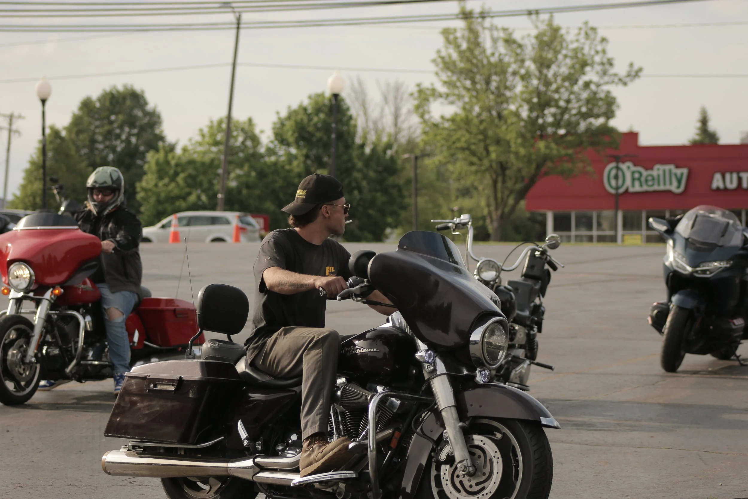 Two motorcyclists sitting on their bikes in a parking lot. One is wearing a black helmet and jacket, the other has a black cap and sunglasses. There are trees, cars, and a red O'Reilly Auto Parts store in the background.