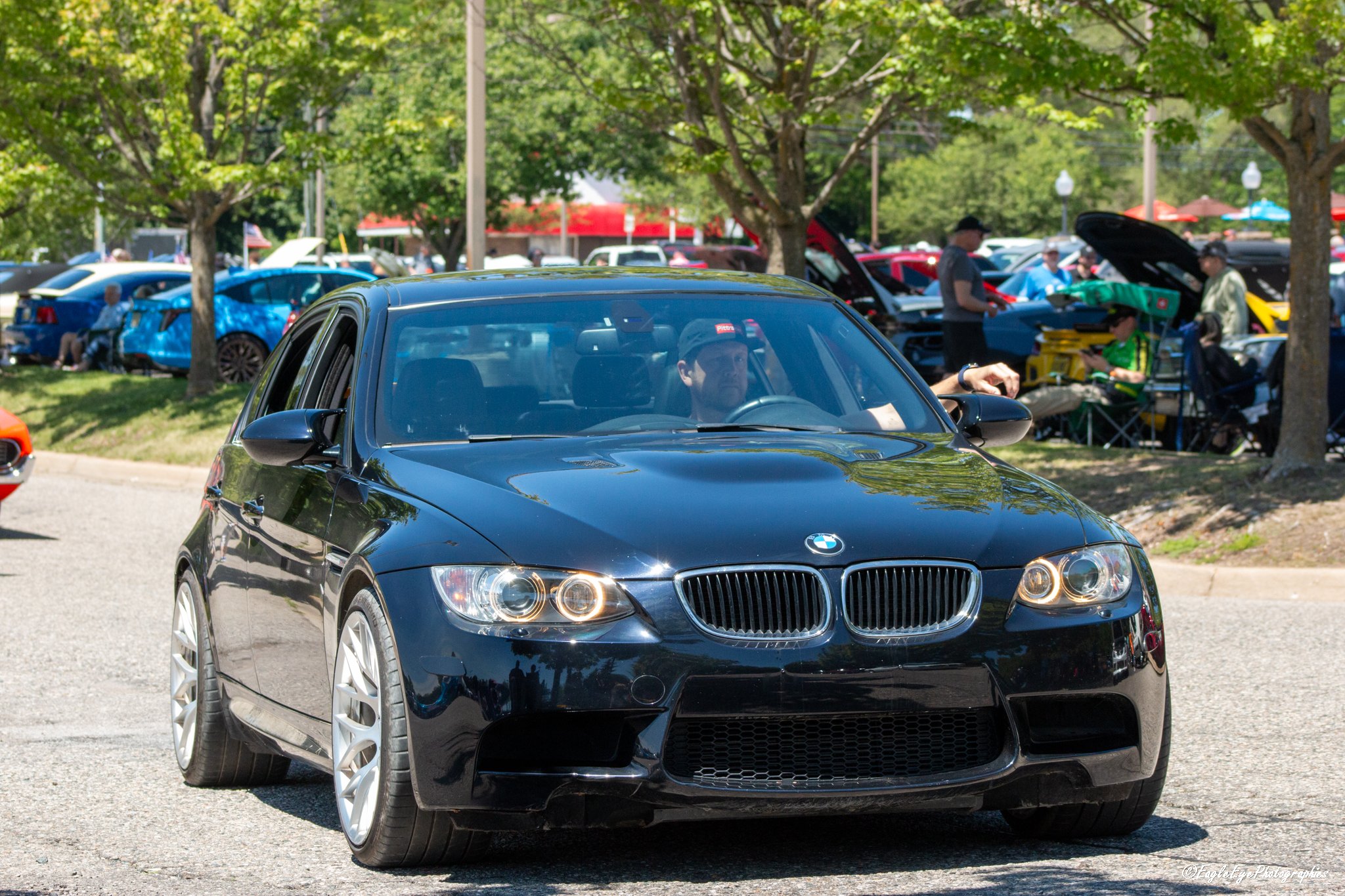 A black BMW coupe parked outdoors at a car show, with other cars and people in the background under trees with green leaves.
