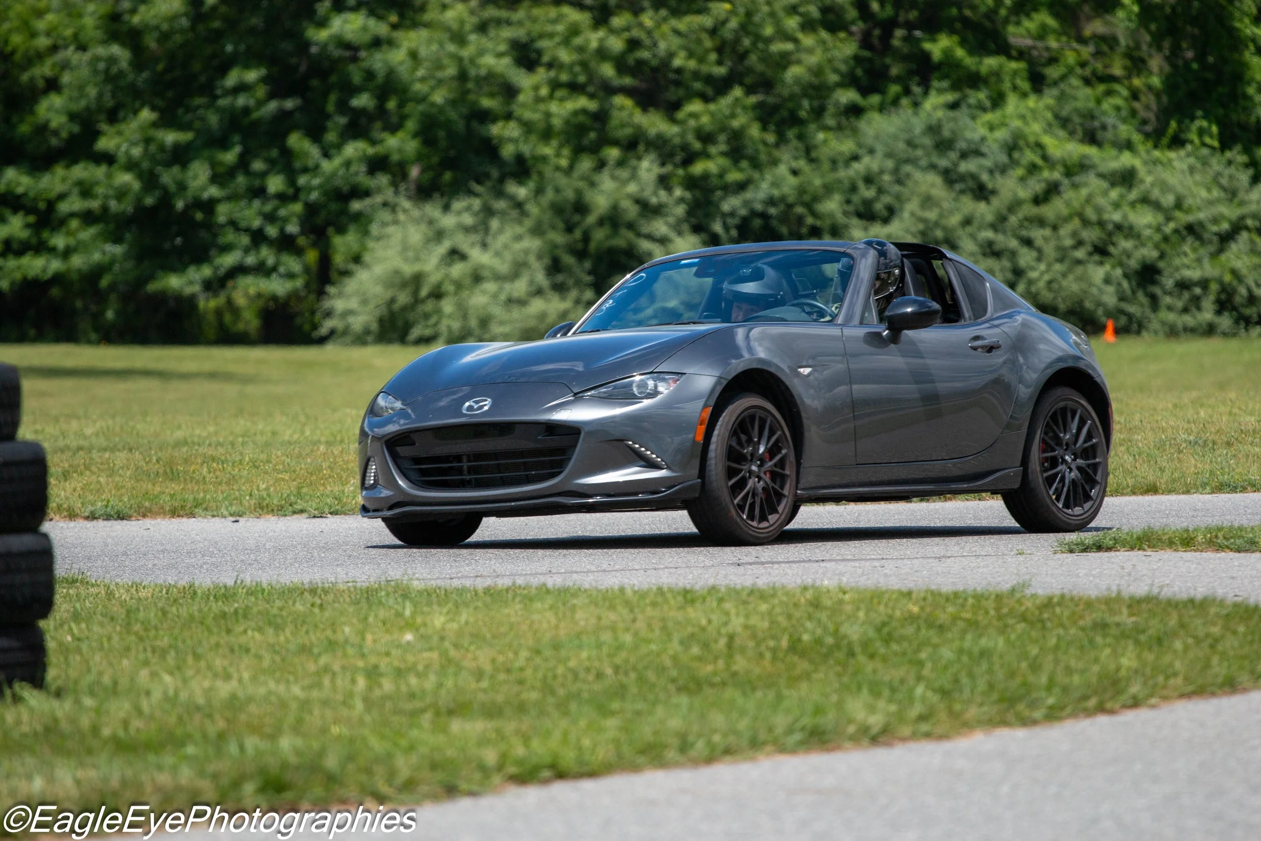 A gray Mazda convertible car parked on grass with green trees in the background.