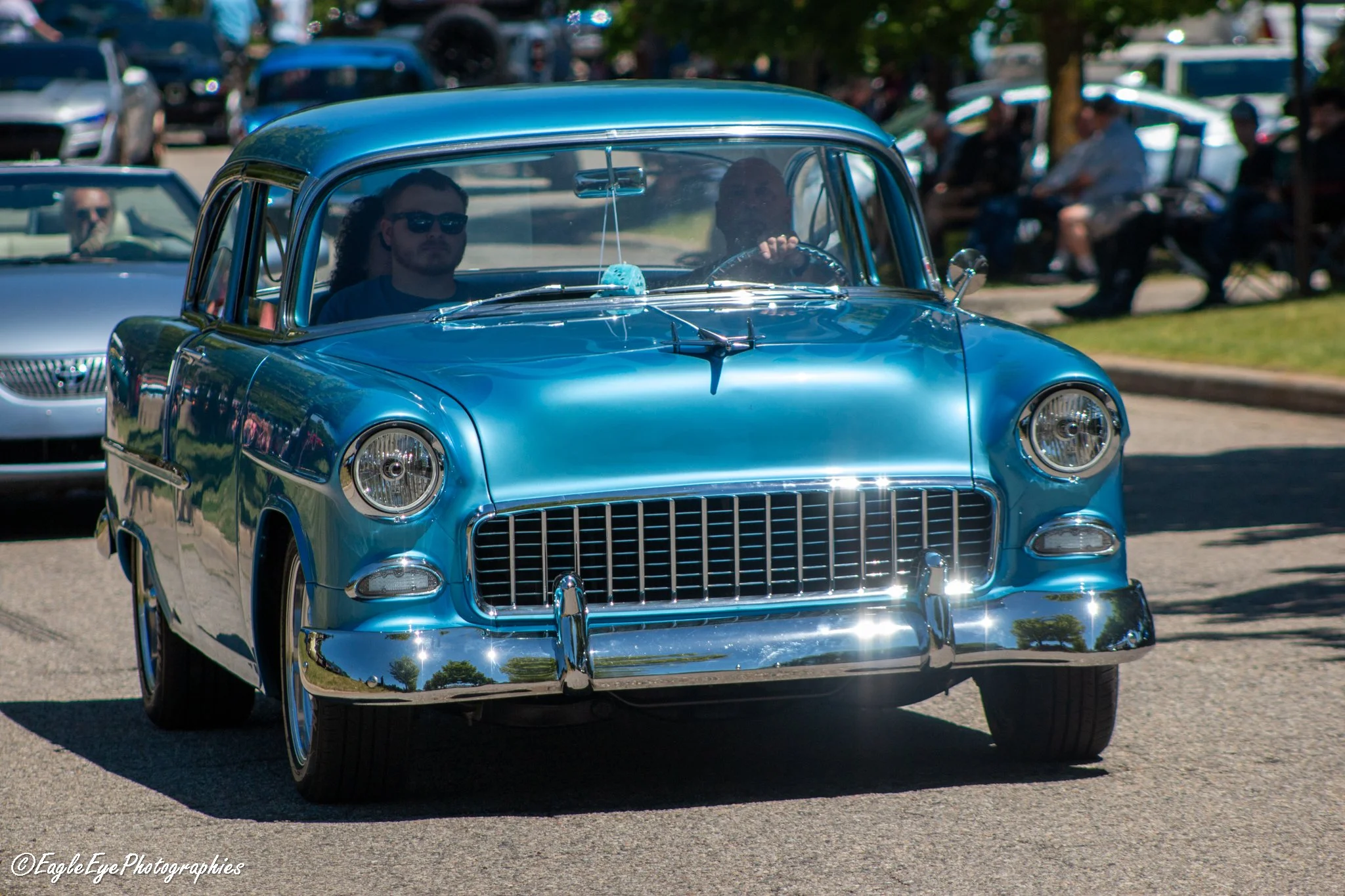 A vintage blue car on display at a car show with people sitting on benches in the background.