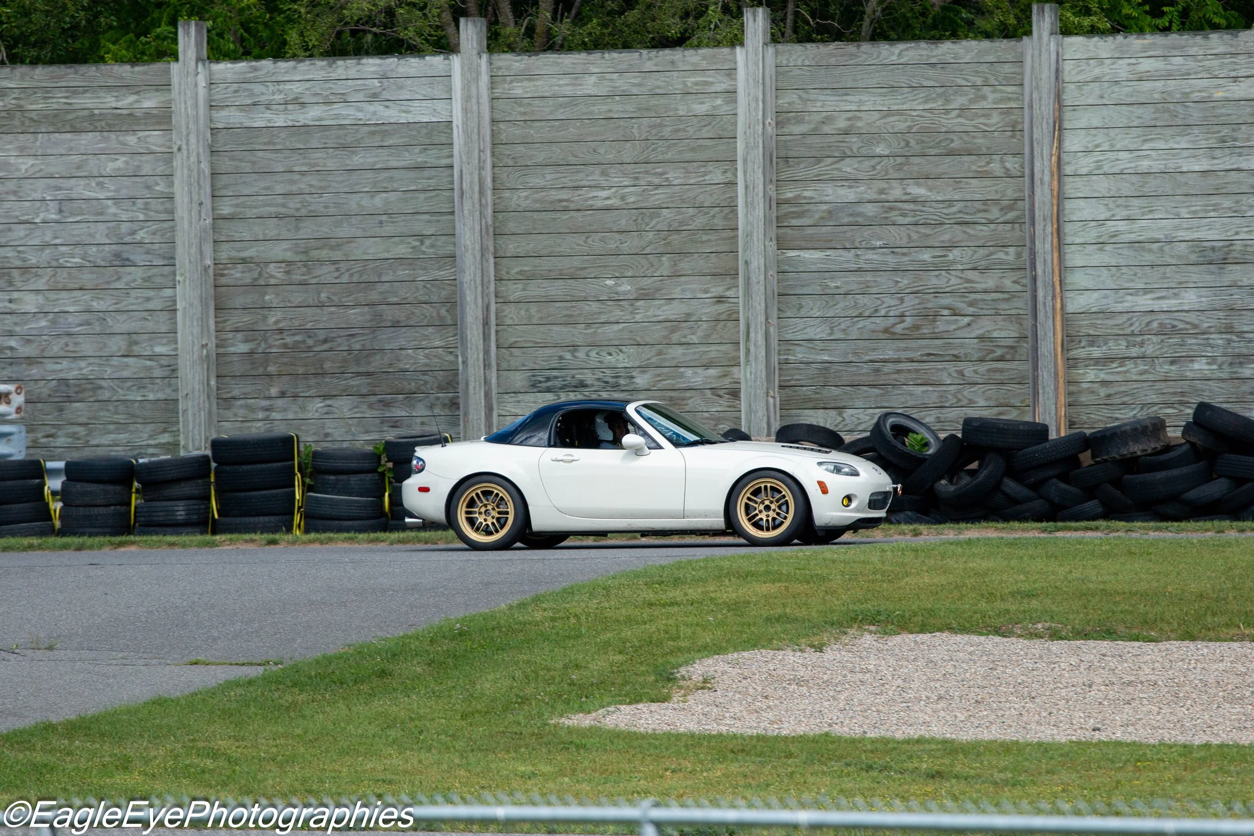 A white convertible sports car with gold wheels parked in front of a wooden fence, surrounded by used tires stacked along the fence line.