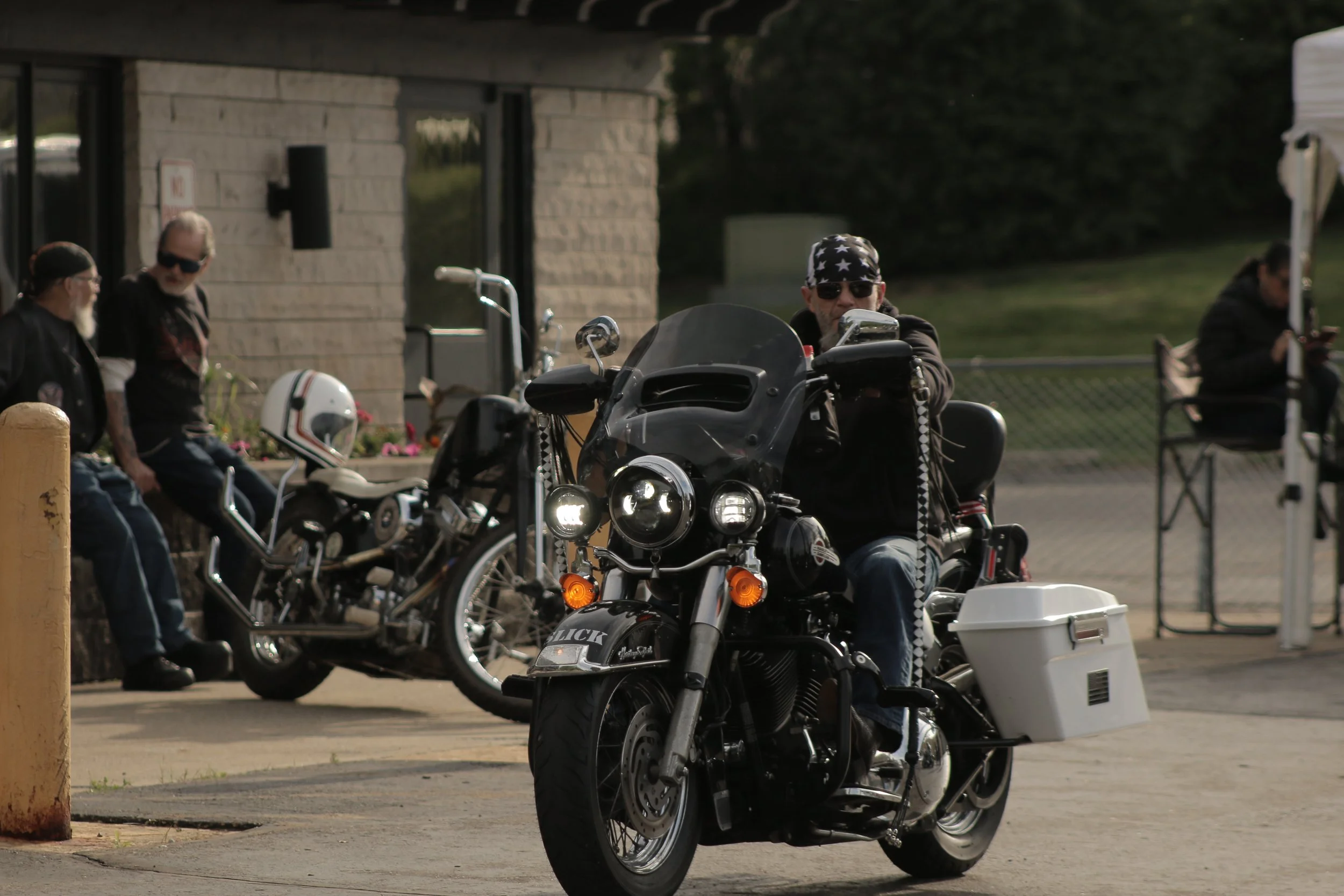 Man wearing sunglasses and bandana sitting on a black motorcycle with white storage compartment, parked in a lot with other motorcycles and people standing nearby.