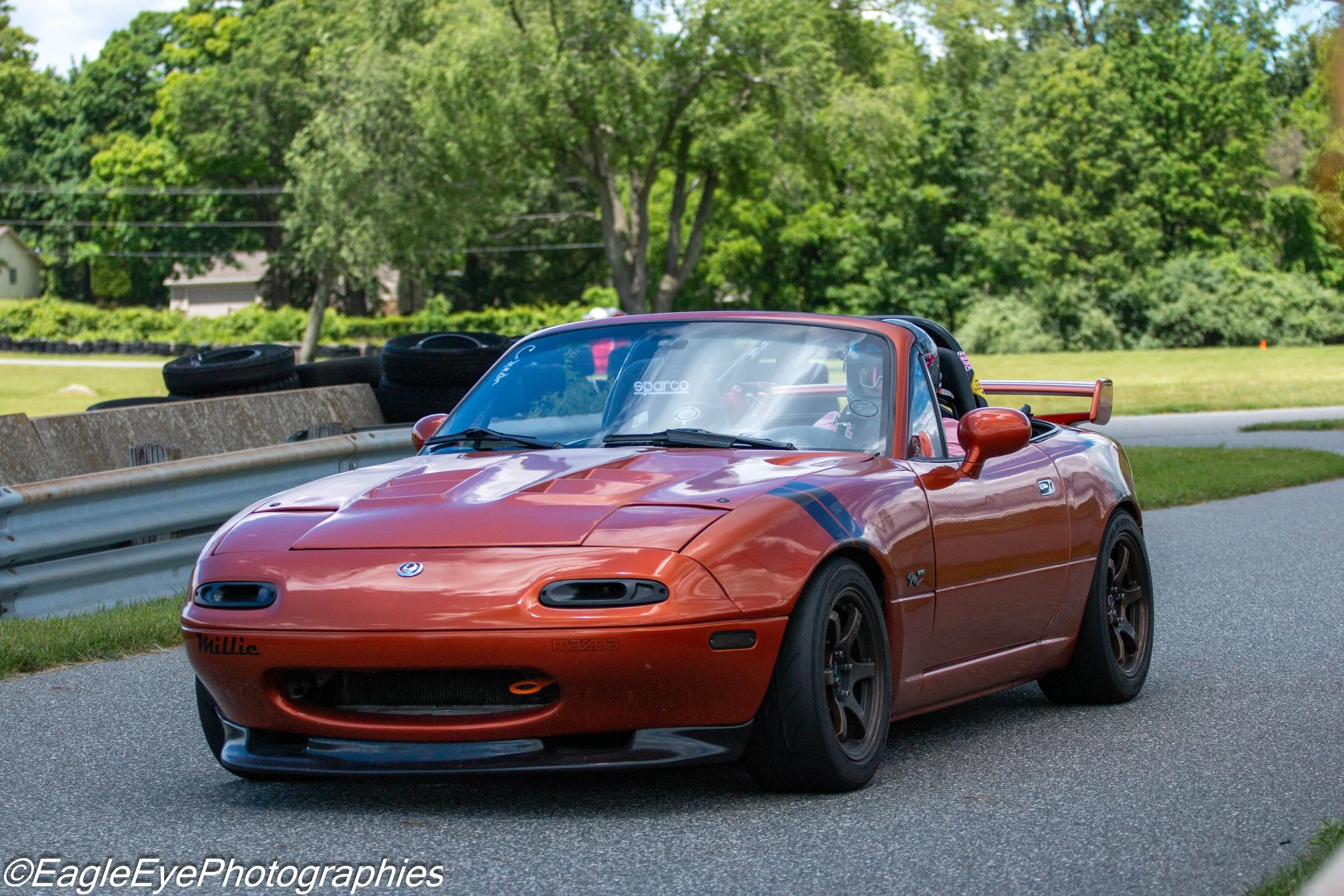 An orange Mazda MX-5 Miata sports car driving on a road next to a grassy area with trees in the background.