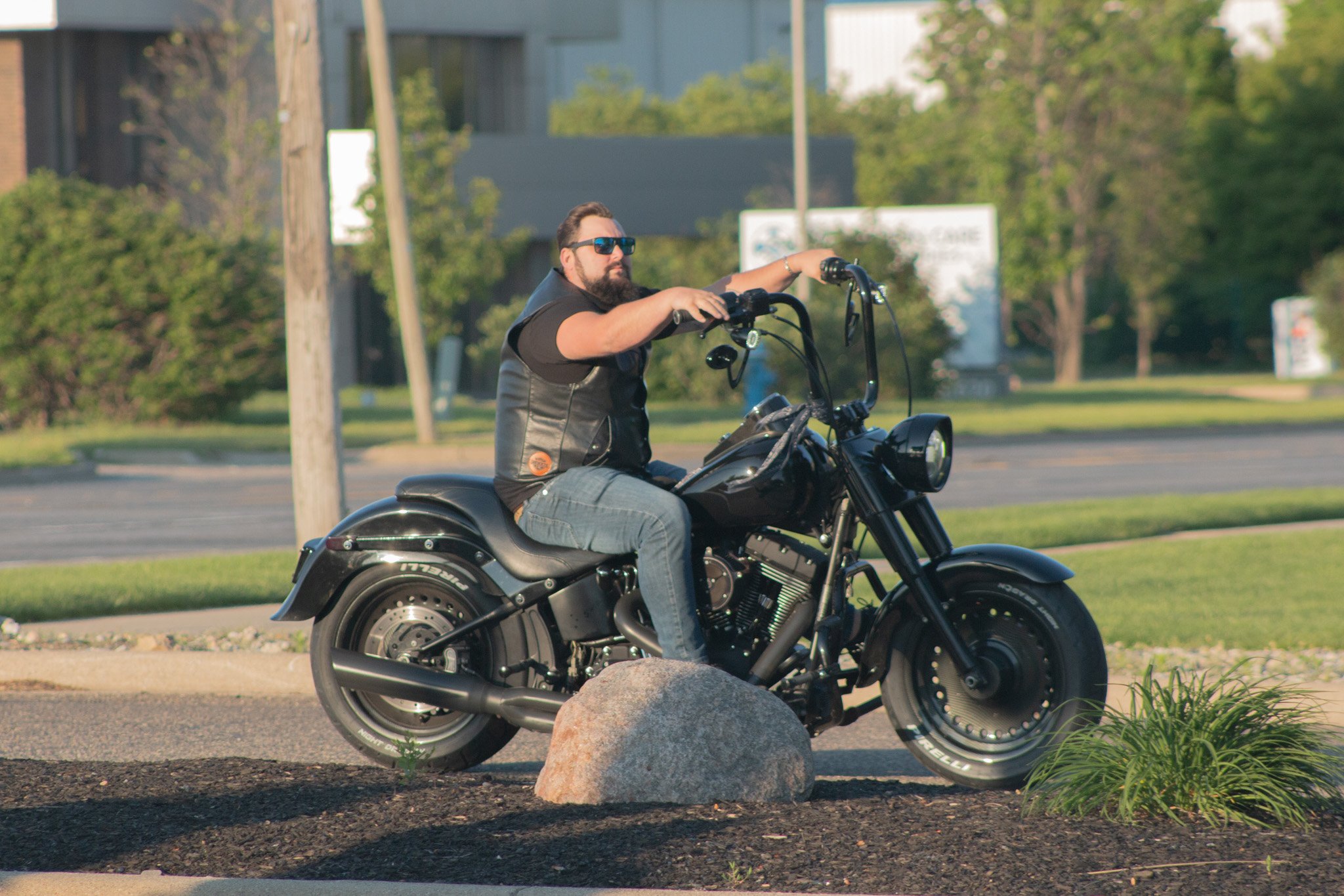 Man riding a black motorcycle on a street, wearing sunglasses, a black vest, and jeans, with trees and buildings in the background.
