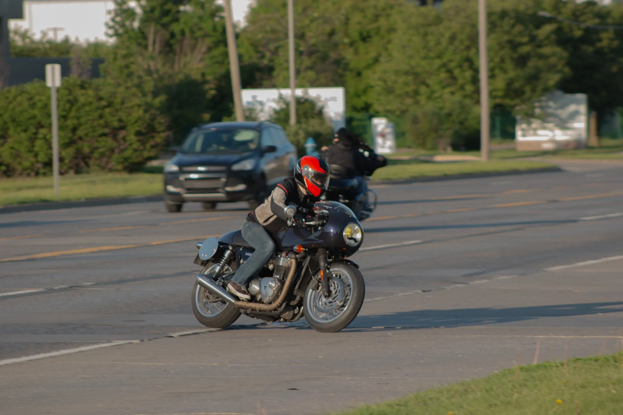 Motorcycle rider wearing a helmet and black jacket riding on a paved road with cars and greenery in the background.