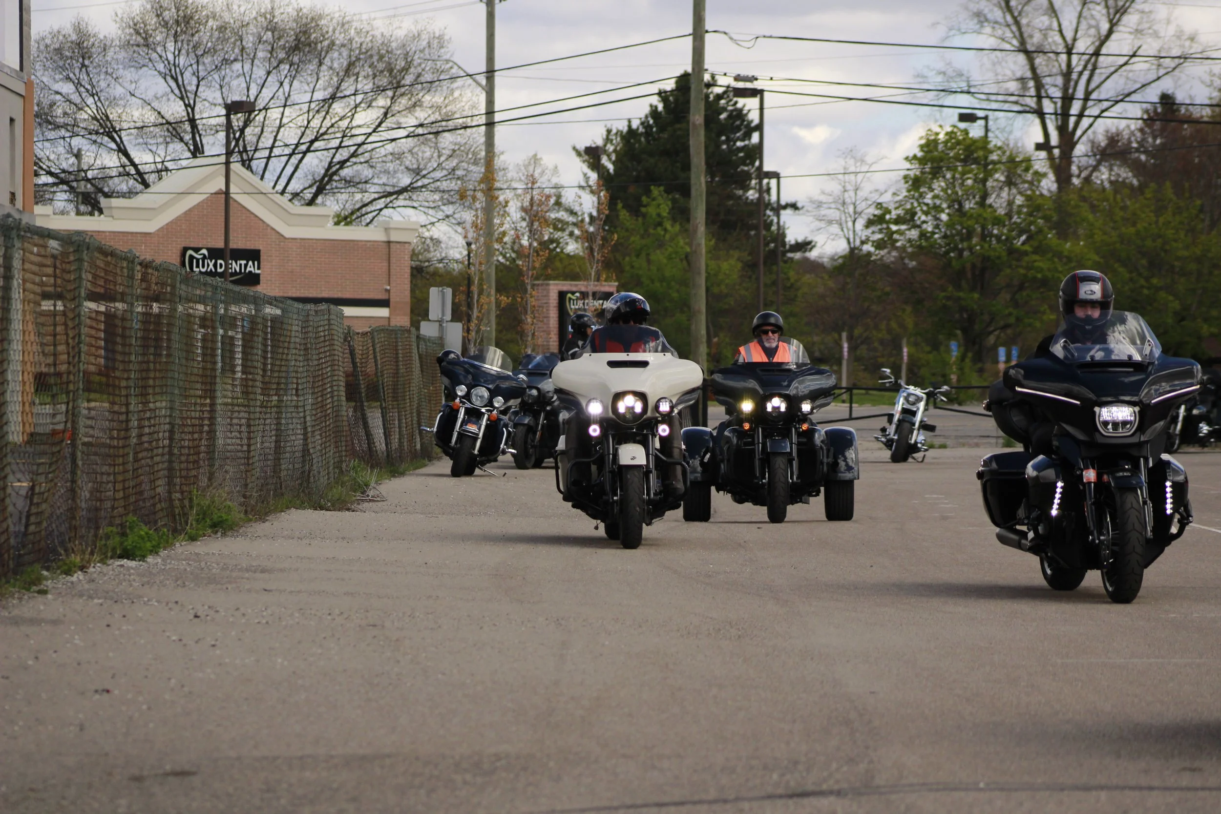 A group of motorcyclists riding down a street in front of a brick building with a sign that says 'Lux Dental'. There is a fence on the left side and power lines overhead. Trees with leafless branches are visible in the background.