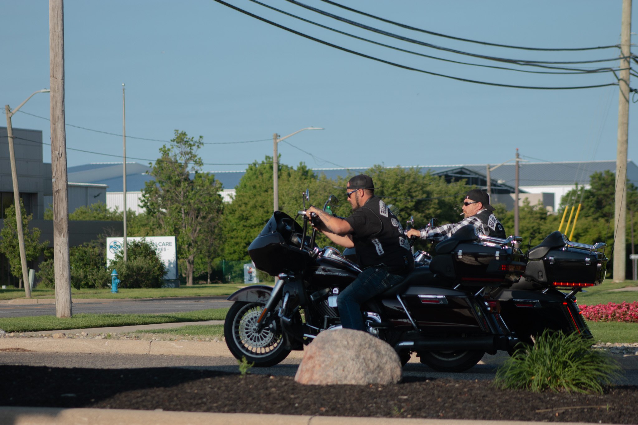 Two motorcyclists riding on a road with trees, buildings, power lines, and a fire hydrant visible in the background.