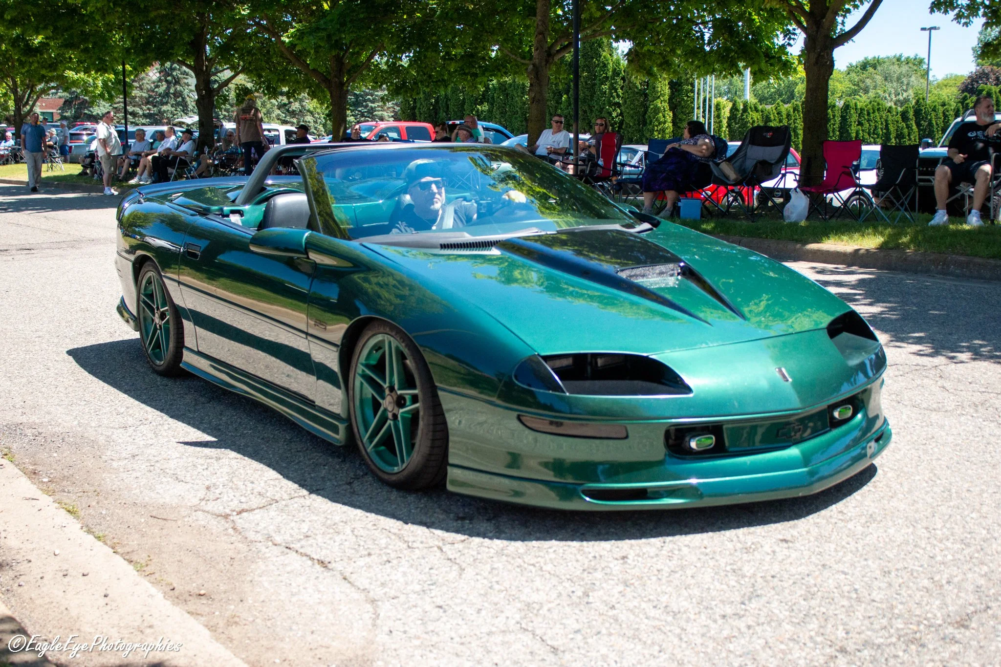 A teal sports car with a convertible top parked on a street beneath green leafy trees during an outdoor event, with people gathered in the background.