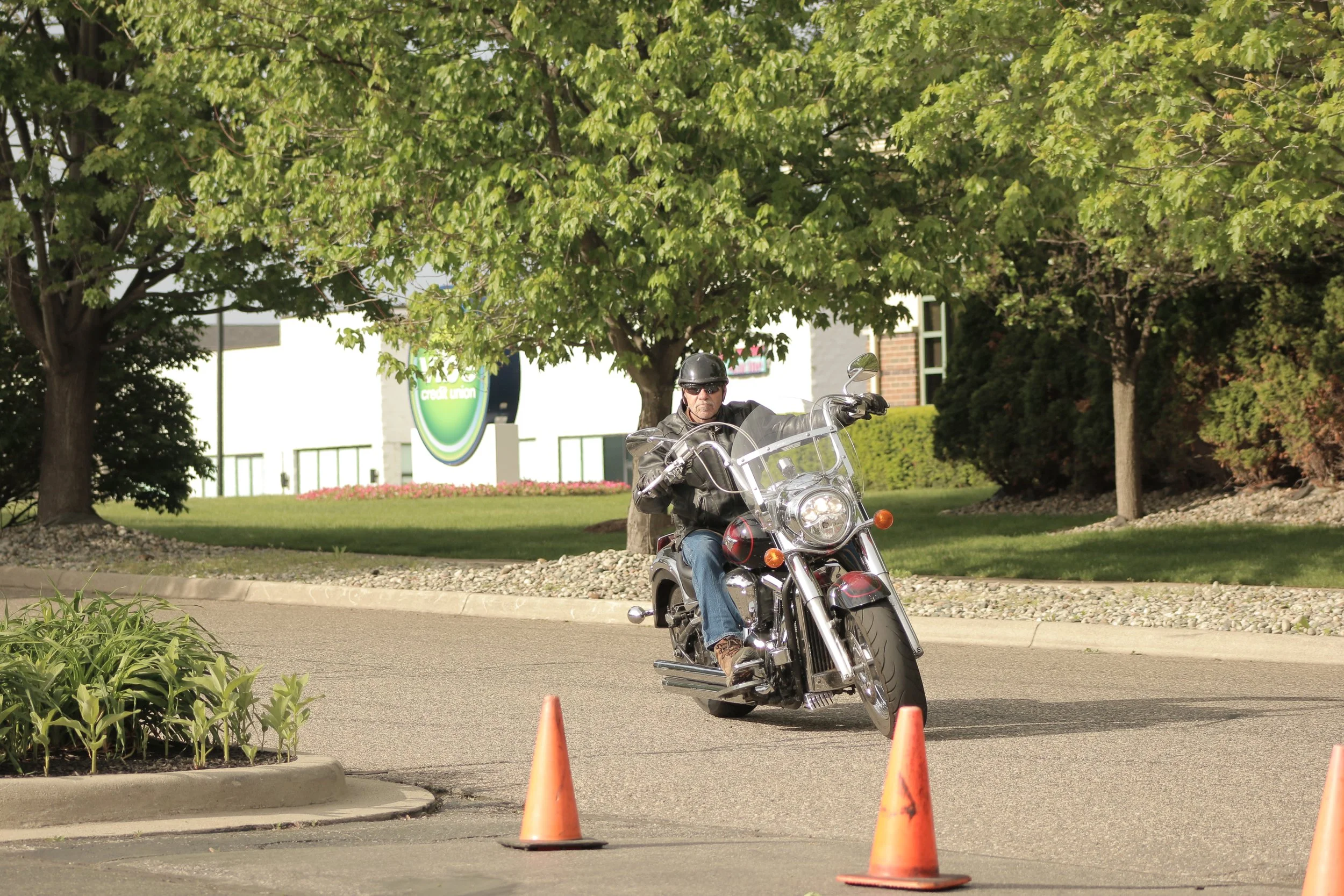 A man riding a motorcycle between two orange traffic cones on a street with trees and a white building in the background.