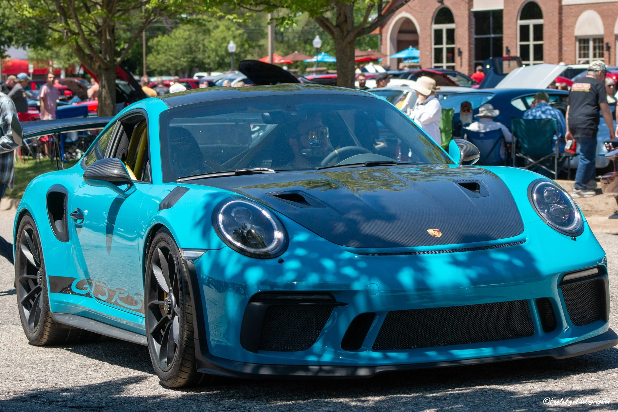 A bright blue sports car, likely a Porsche, on display at an outdoor car show with other cars and people in the background.