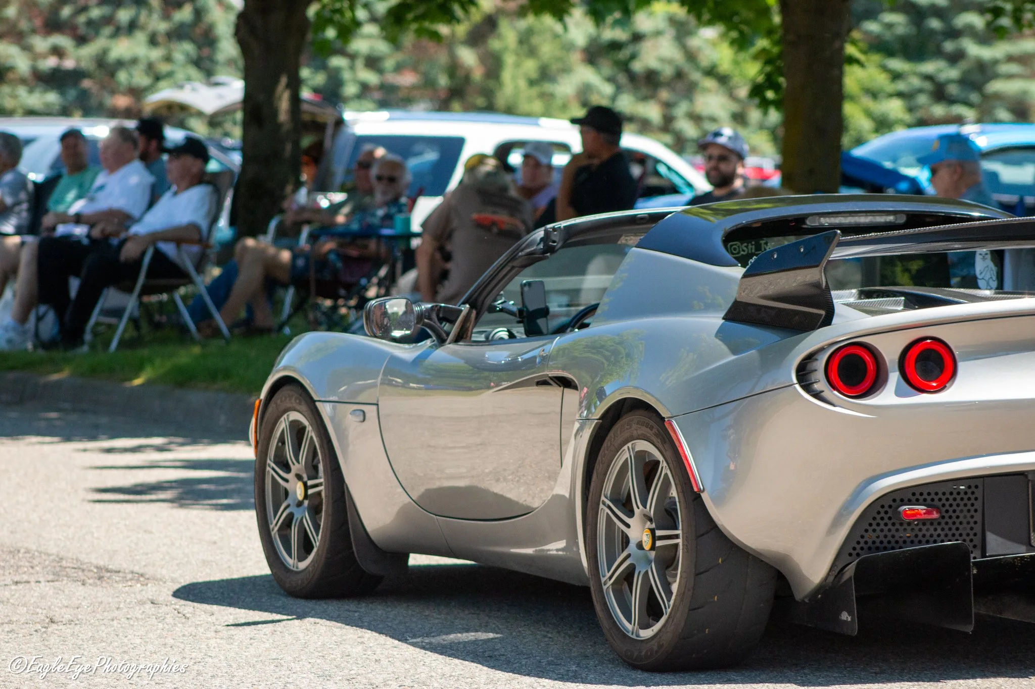 A silver sports car with a rear spoiler parked in front of a group of people sitting under trees at an outdoor event, with parked cars in the background.