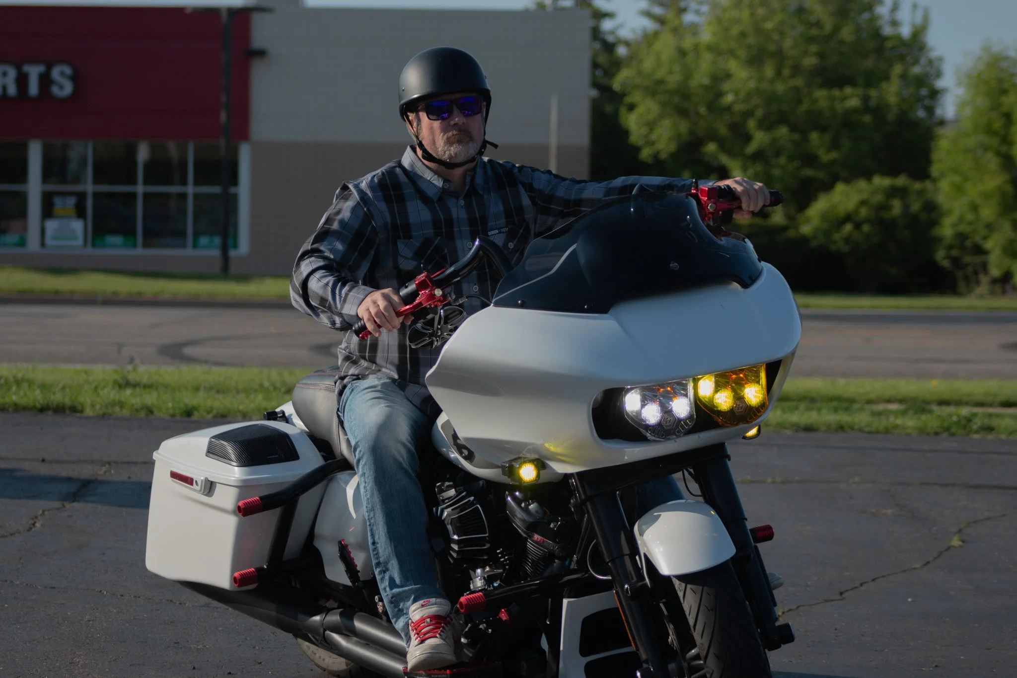 A man riding a white motorcycle with a black front windscreen, wearing a black helmet, sunglasses, a plaid shirt, and jeans, in a parking lot with trees and a building in the background.