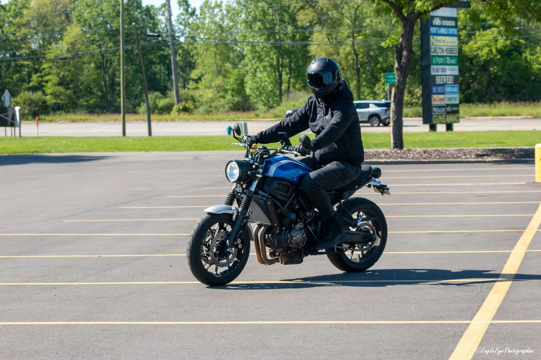 A person riding a black motorcycle in a parking lot, wearing a black helmet and jacket, with greenery and trees in the background.