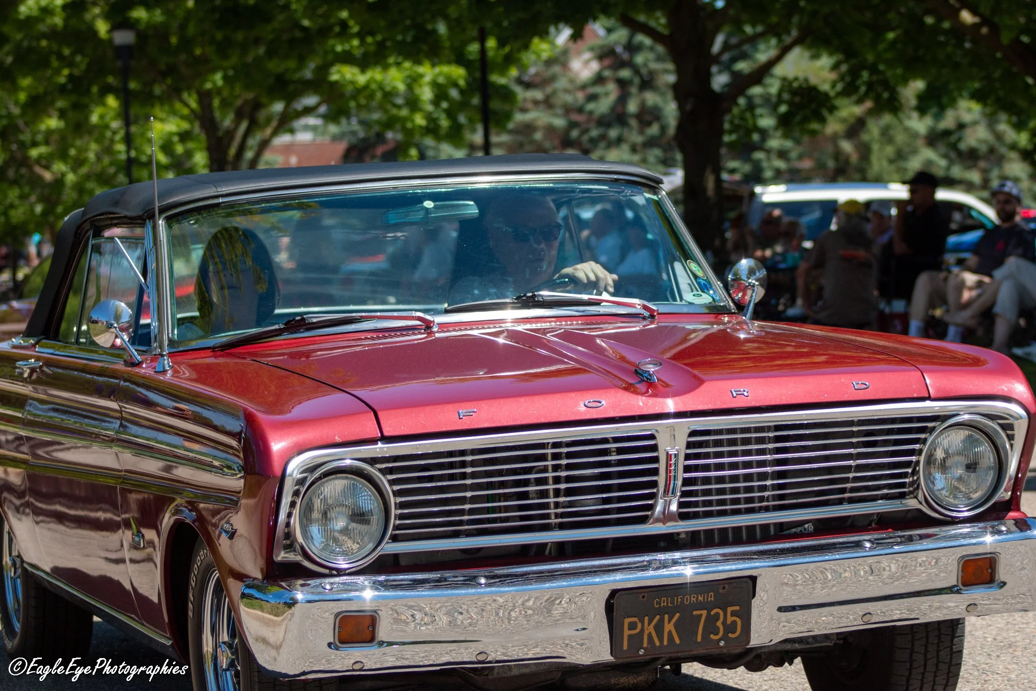 A vintage red Ford convertible with a black soft top parked outdoors during daylight, with people in the background under trees.