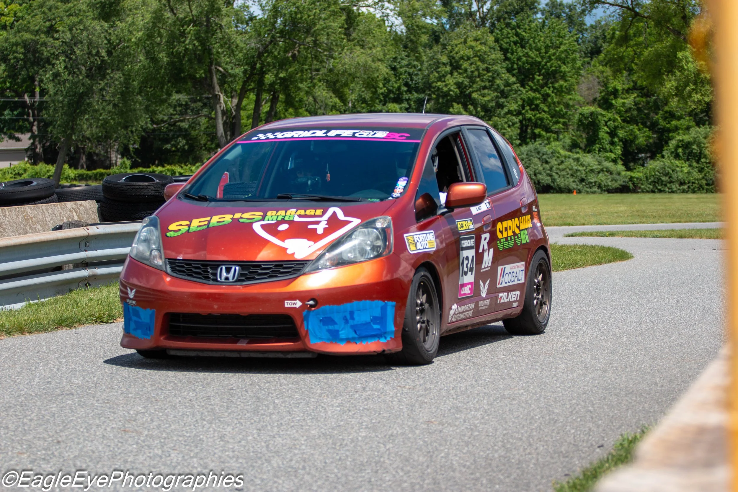 Red Honda racing car with various sponsor decals, driving on a race track surrounded by green trees and tires stacked along the side.