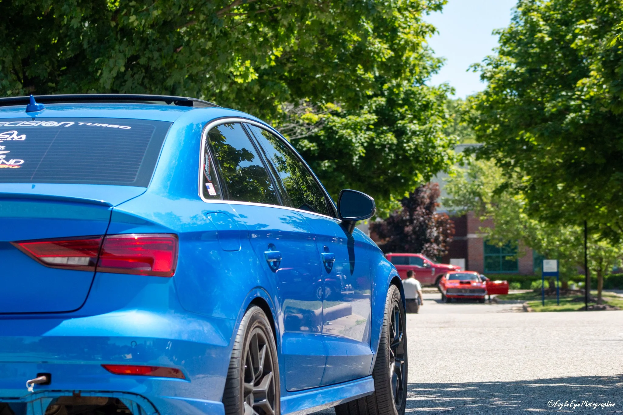 A blue sports car parked in a parking lot, with green trees and other cars in the background.