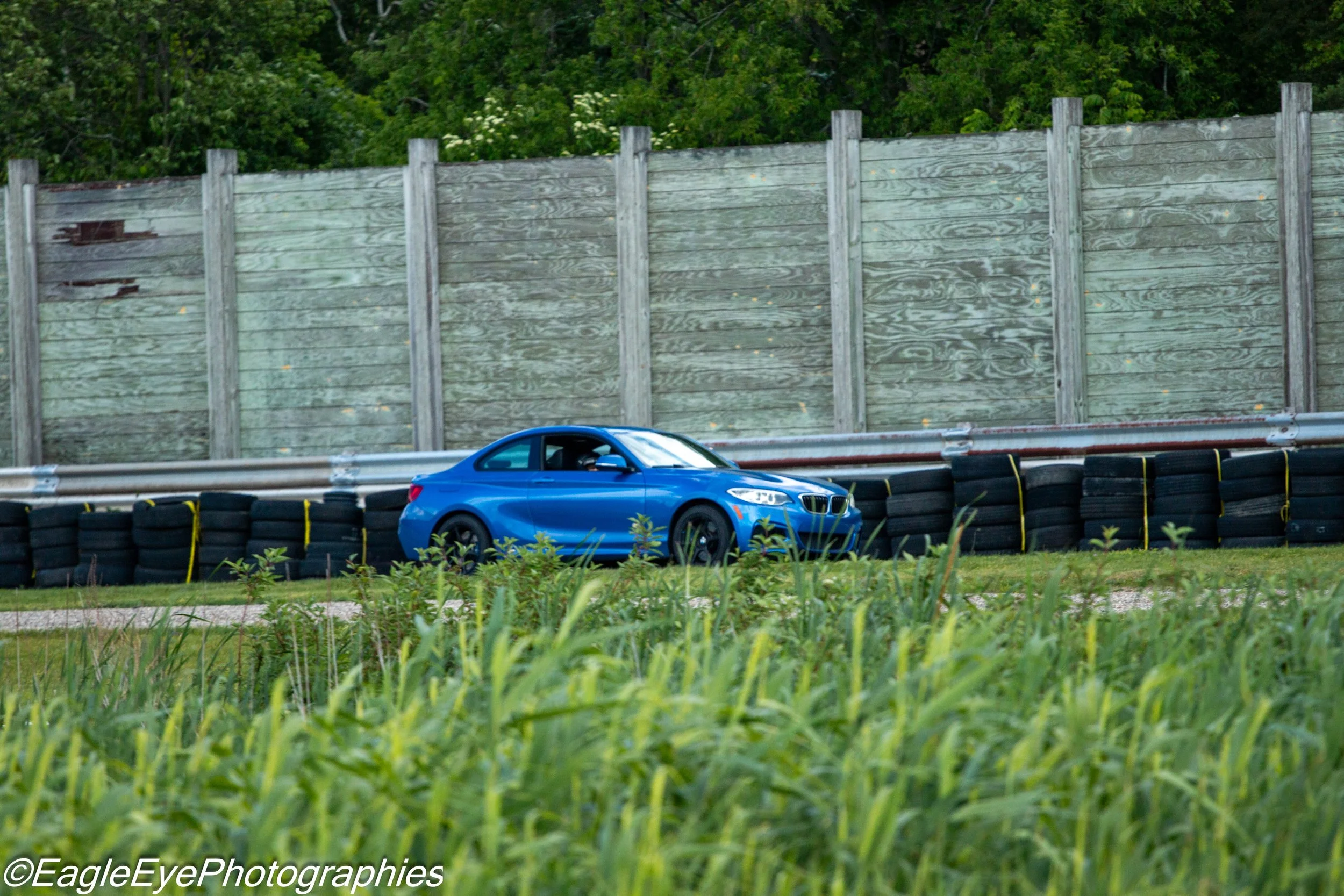 A blue sports car on a race track with smoke behind it, set against a wooden fence and green trees in the background.