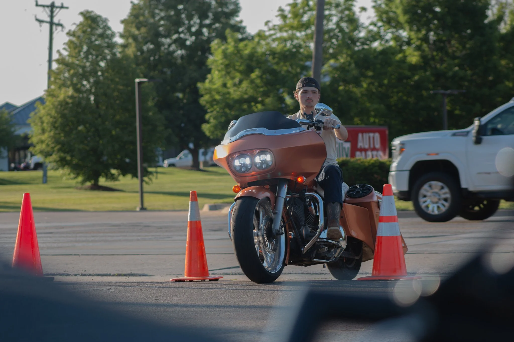 A man rides a motorcycle through orange traffic cones on a paved surface, with trees, a white vehicle, and a sign in the background.
