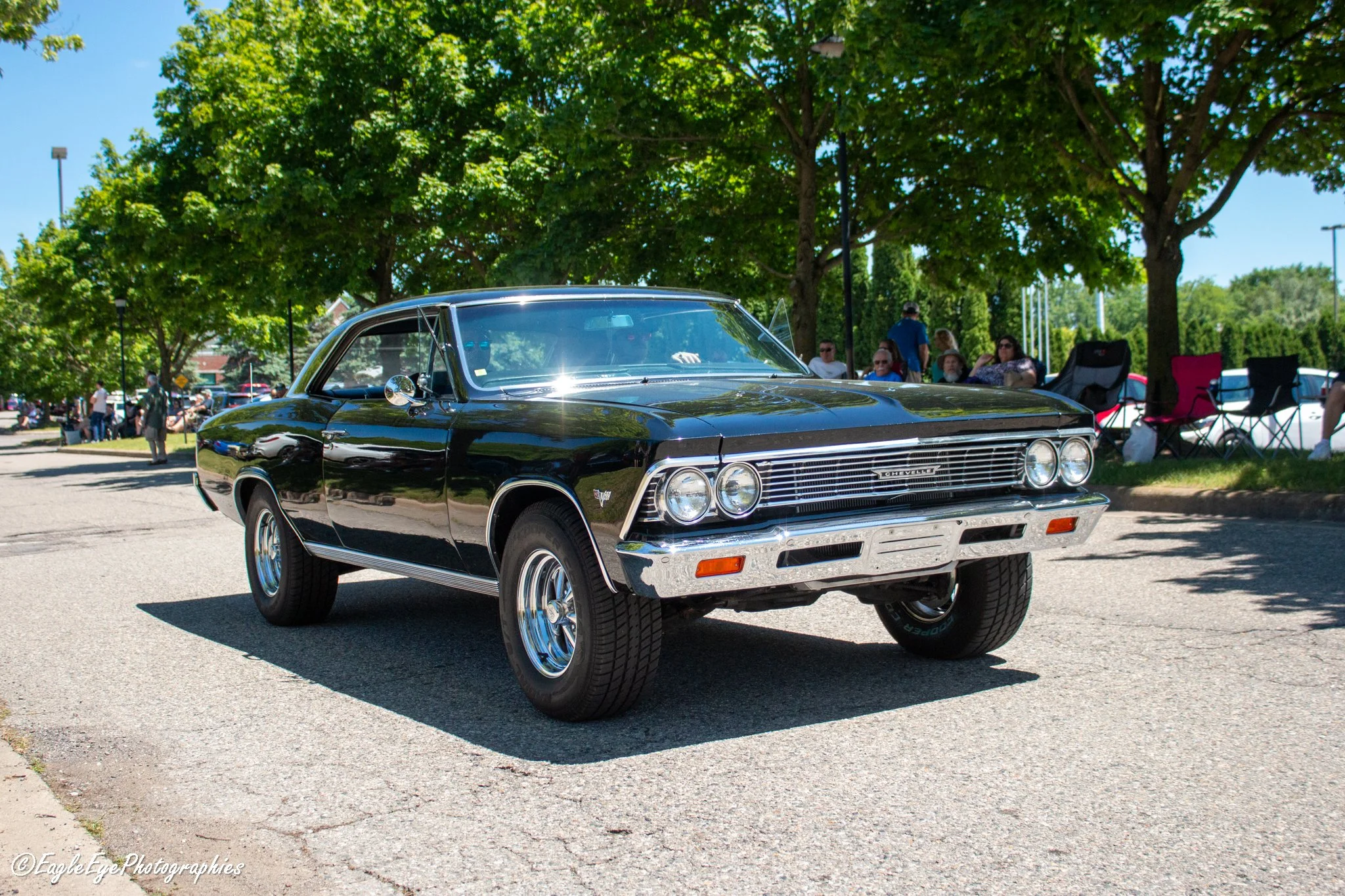 A black vintage Chevrolet car on display outdoors at a car show, with a group of people and trees in the background.