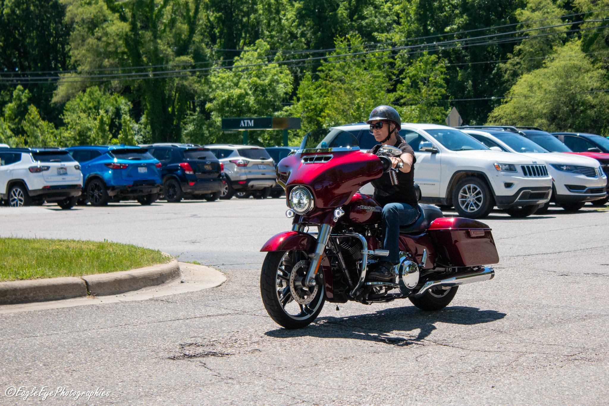 A man wearing sunglasses, a black helmet, and a black shirt riding a maroon motorcycle in a parking lot with several parked cars and green trees in the background.