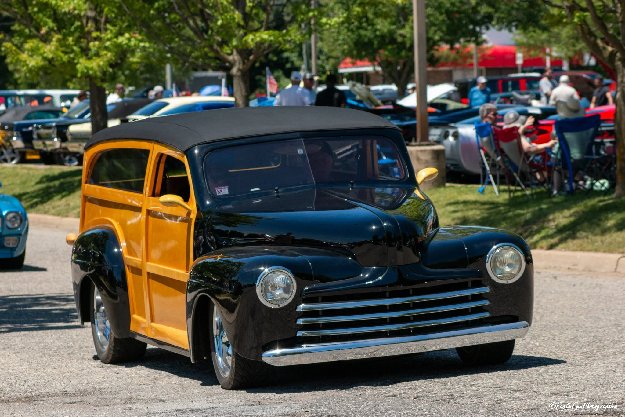 A vintage yellow and black car with a convertible top parked outdoors during an event, with other cars and people visible in the background under trees.