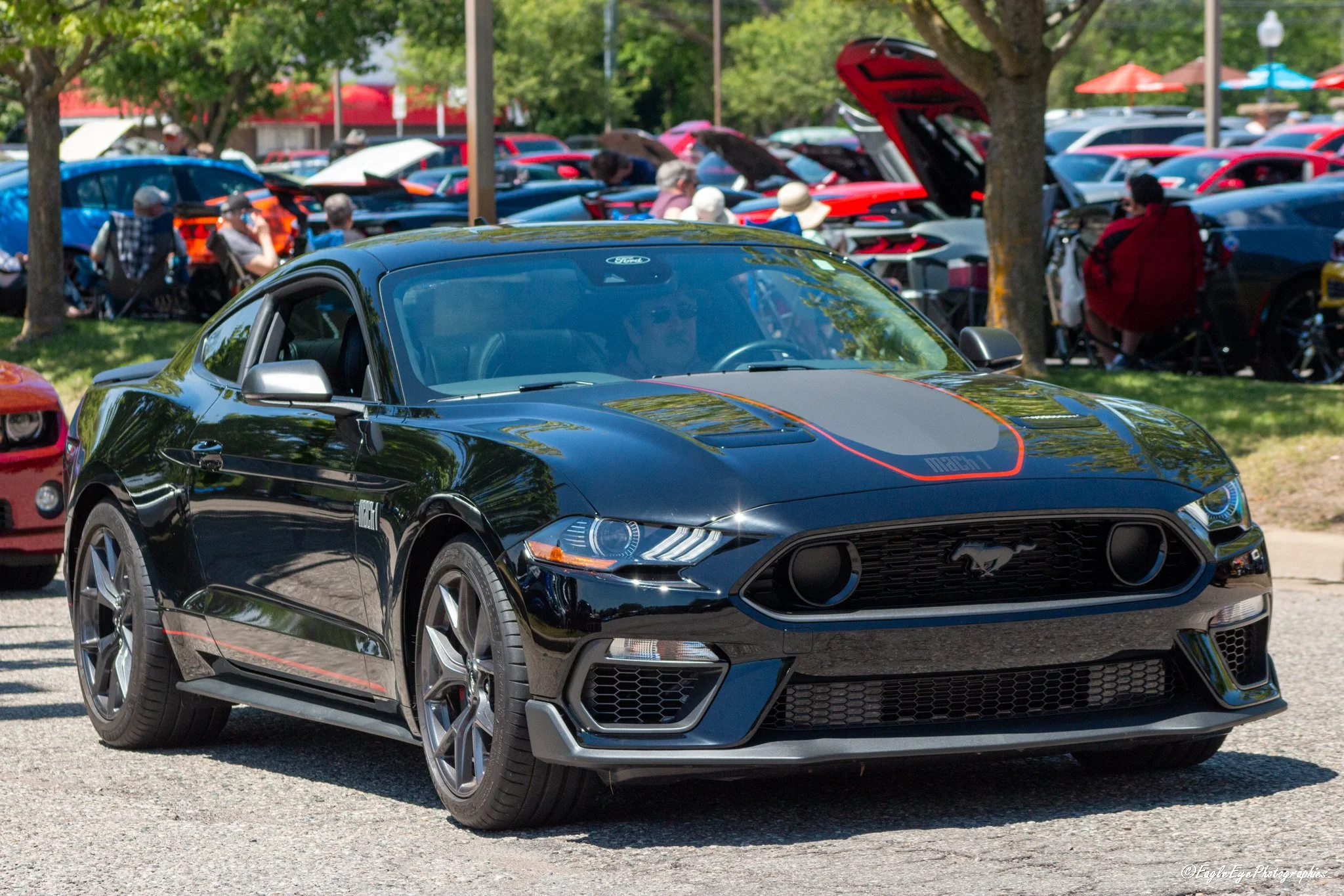 A black Ford Mustang sports car parked at an outdoor car show, with other cars and people in the background.