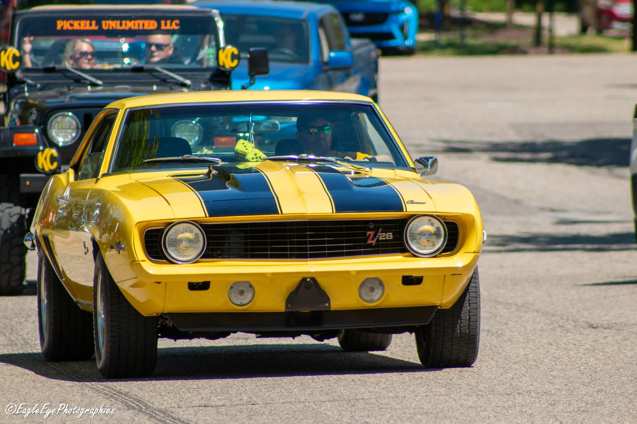 A yellow Chevrolet Camaro Z28 driving on a paved surface, with a black stripe running down the center of the hood. A man with sunglasses is driving, and a dice decoration hangs from the rearview mirror. In the background, there are several other vehi
