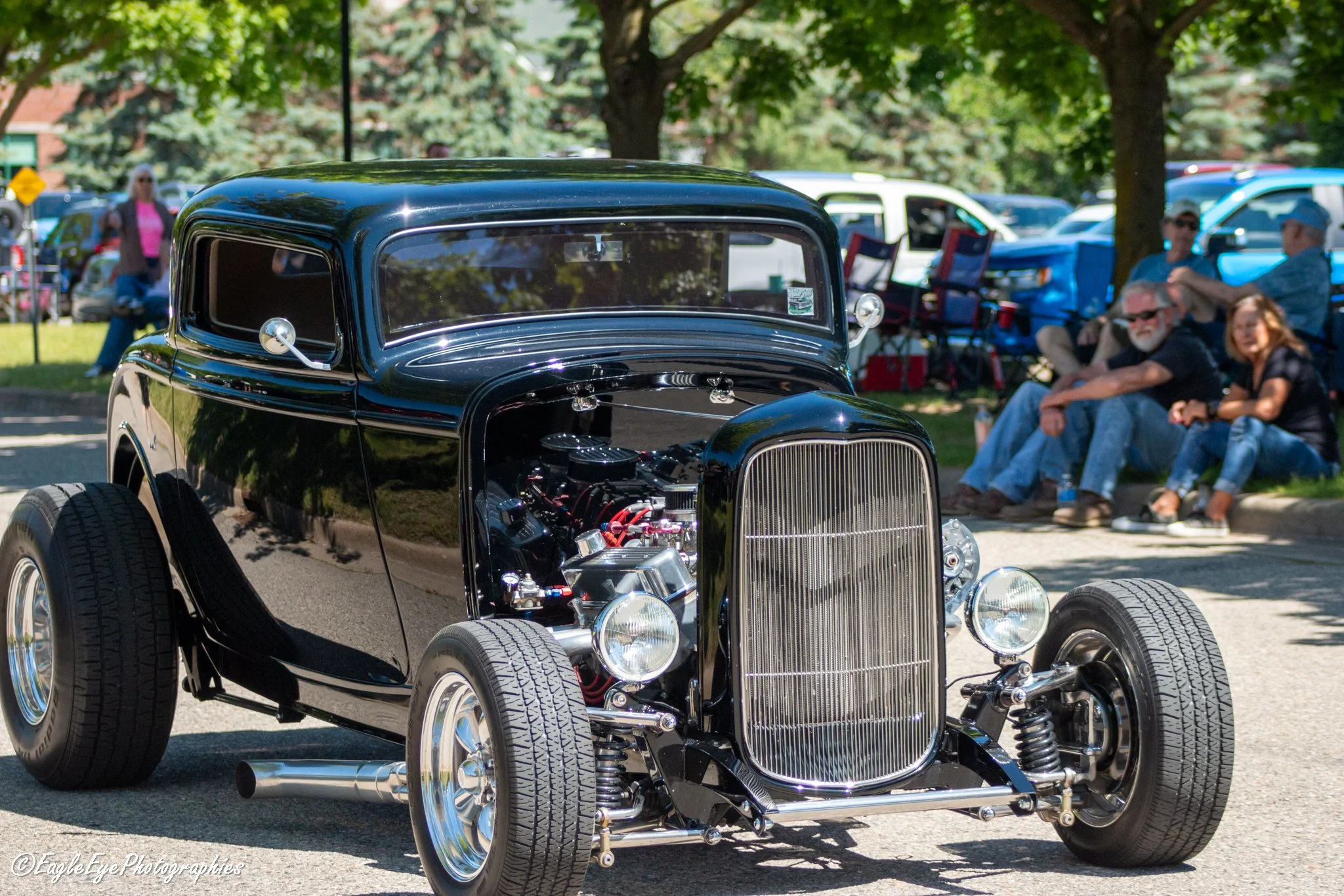 Close-up of a vintage black car with shiny chrome details at an outdoor car show, with people seated and walking in the background under trees.