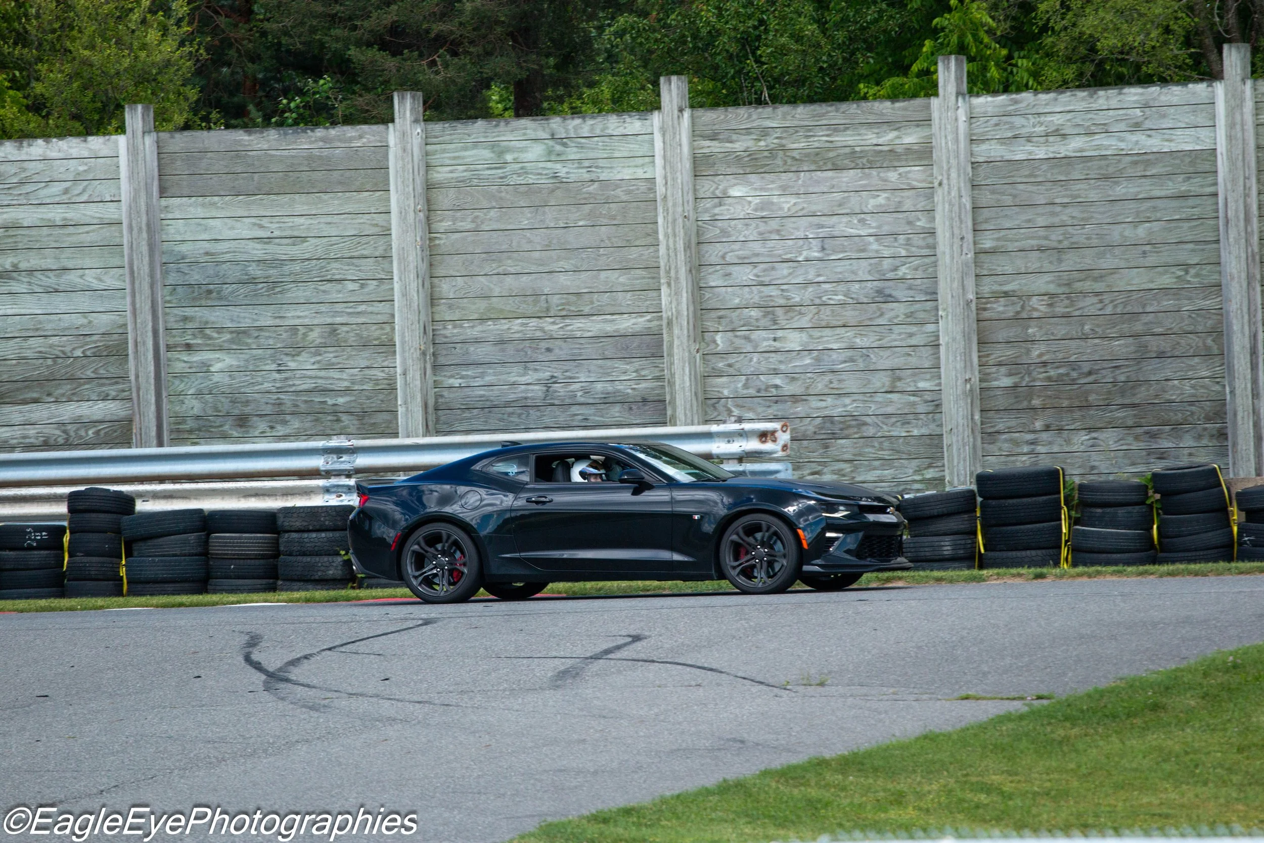 A black sports car driving on a race track with a wall of tires and a tall wooden fence in the background.