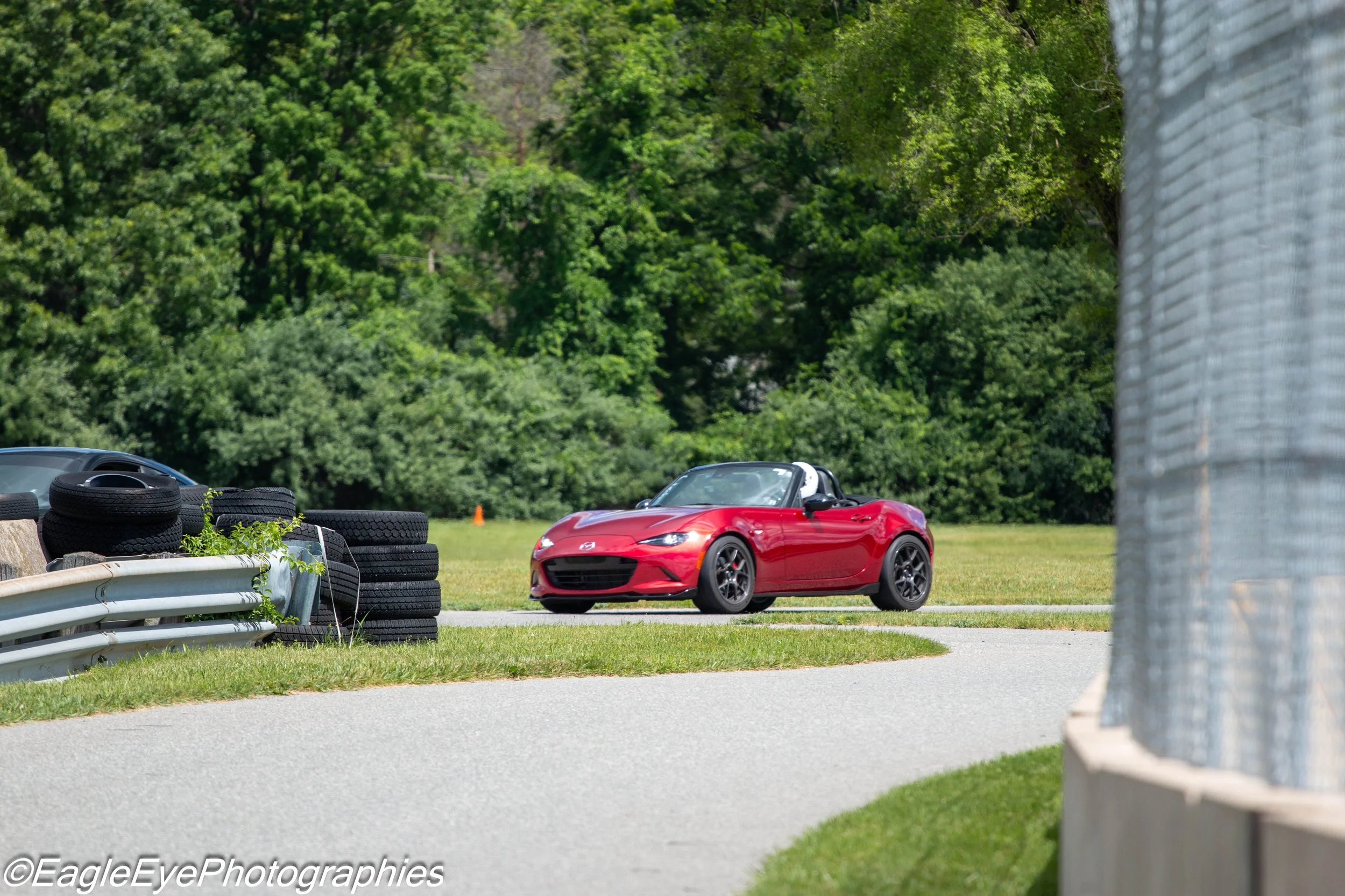 A red Mazda MX-5 Miata convertible car parked on a race track with tires stacked on the side and a metal guardrail in the foreground. Dense green trees are in the background.