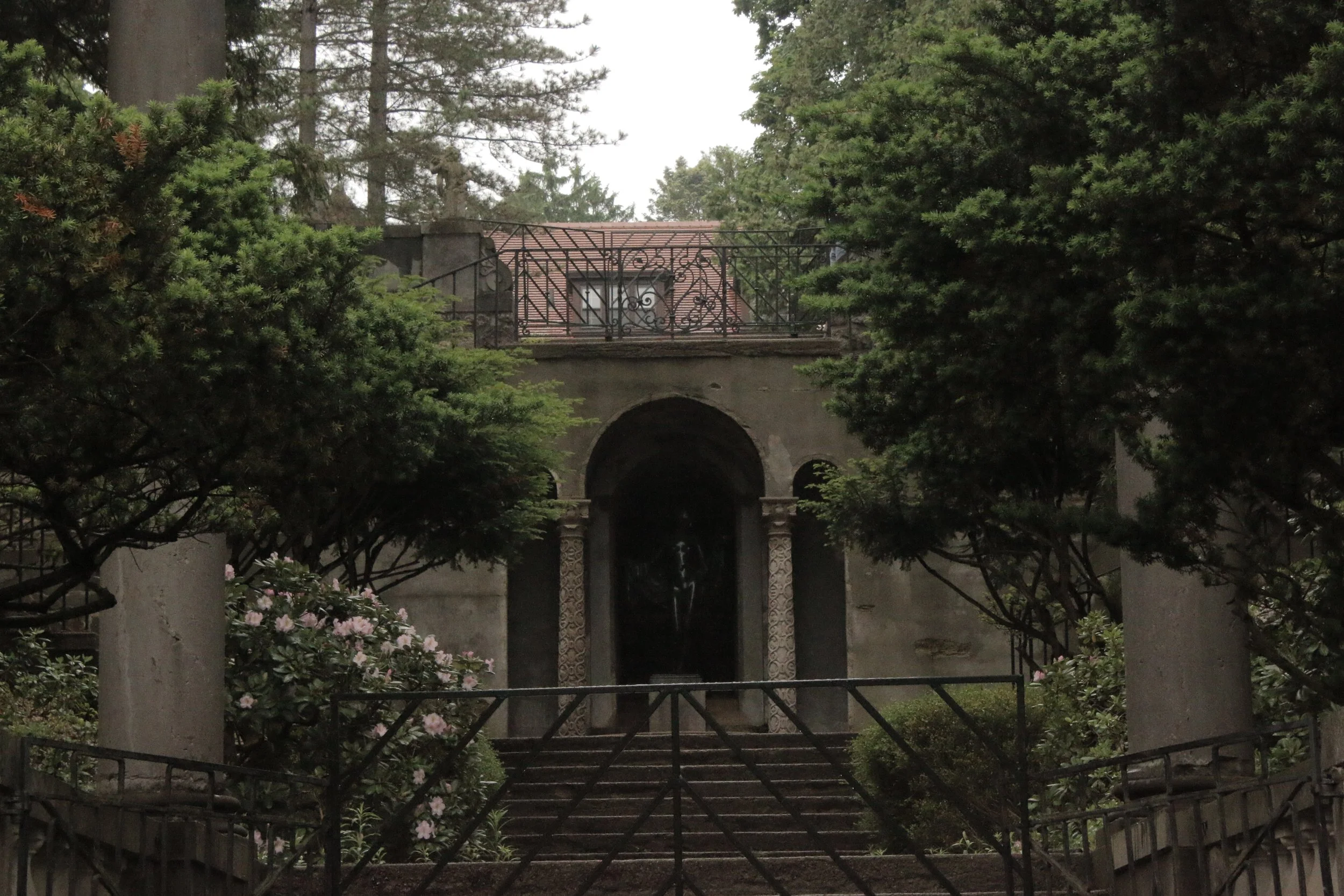 View of a garden with stone archway and wrought iron balcony above, surrounded by lush green trees and pink azalea flowers.