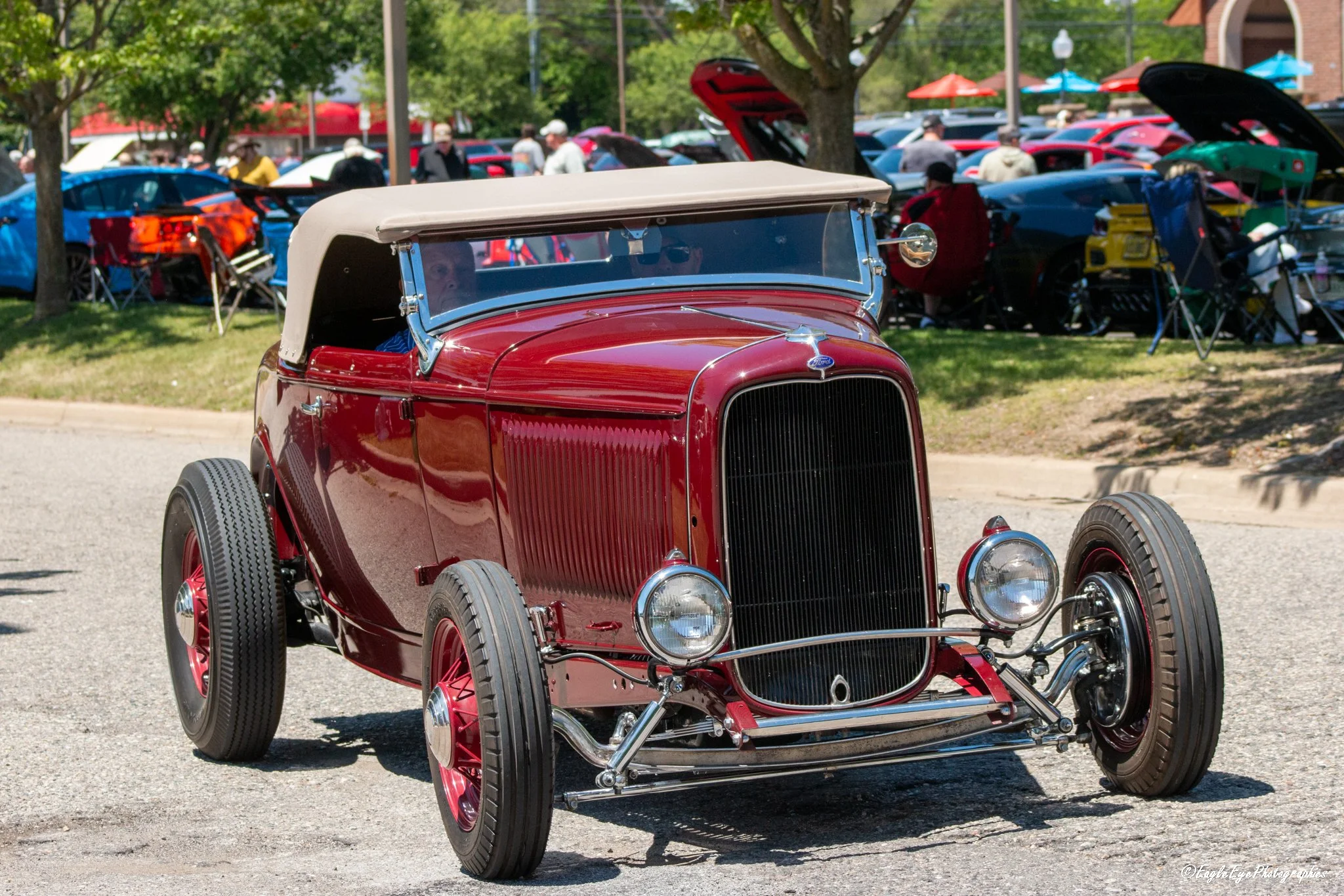 A vintage red car with a beige convertible top parked at a car show with many other cars and people in the background.