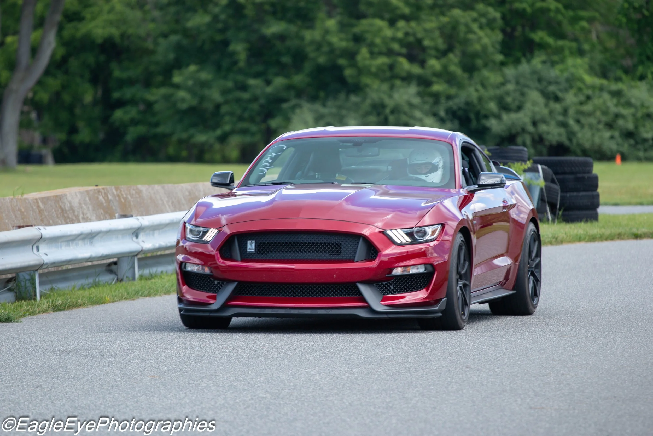 Red sports car with a black grille, driving on a paved road with a guardrail on the side, surrounded by green trees in the background.