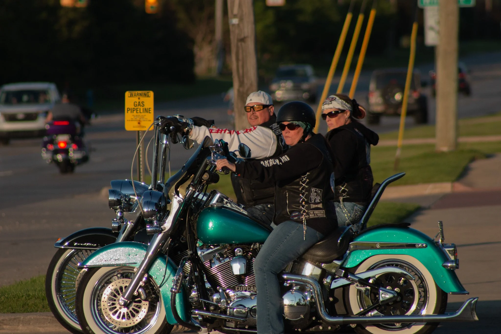 Three motorcyclists, two women and one man, sitting on teal-colored motorcycles in a parking lot during daytime. They are wearing helmets and sunglasses, and appear to be preparing to ride.