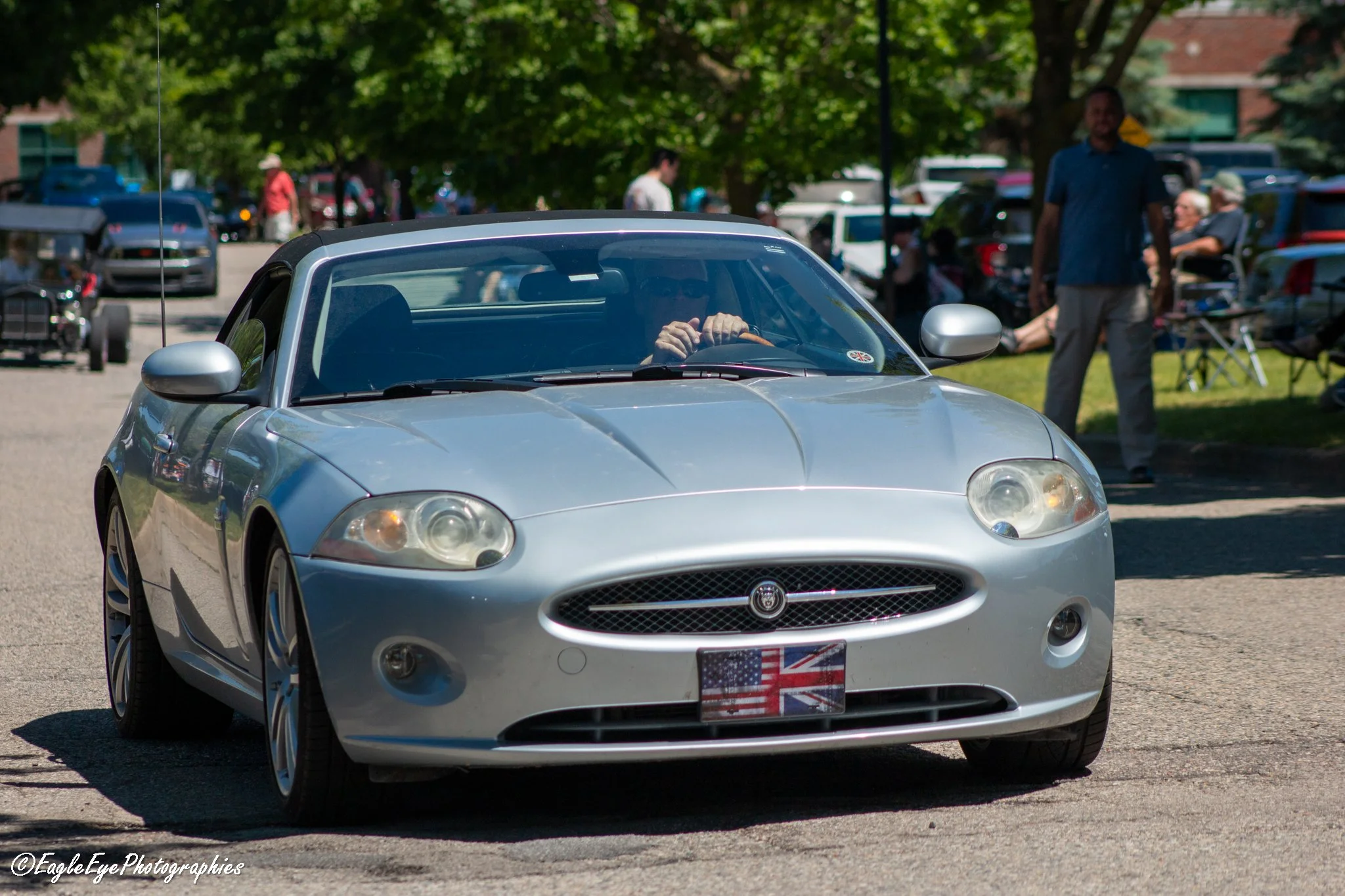A silver convertible sports car with an American flag license plate is parked outdoors on a sunny day, with people and trees in the background.