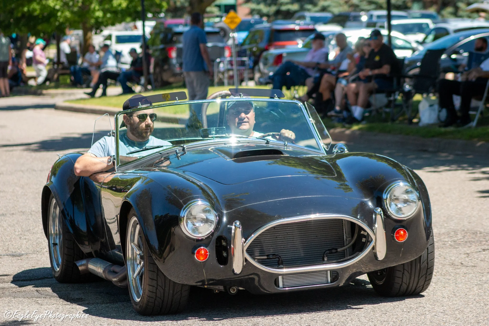 A black vintage sports car with a shiny exterior, driven by a person with short dark hair, sunglasses, and a gray shirt, at an outdoor event on a sunny day. In the background, several people are seated on benches and appear to be watching something, 
