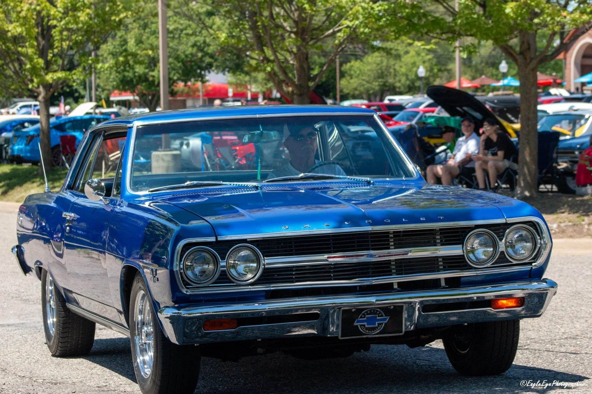 A vintage blue Chevrolet car displayed at a car show, with people sitting under trees and other colorful cars nearby.