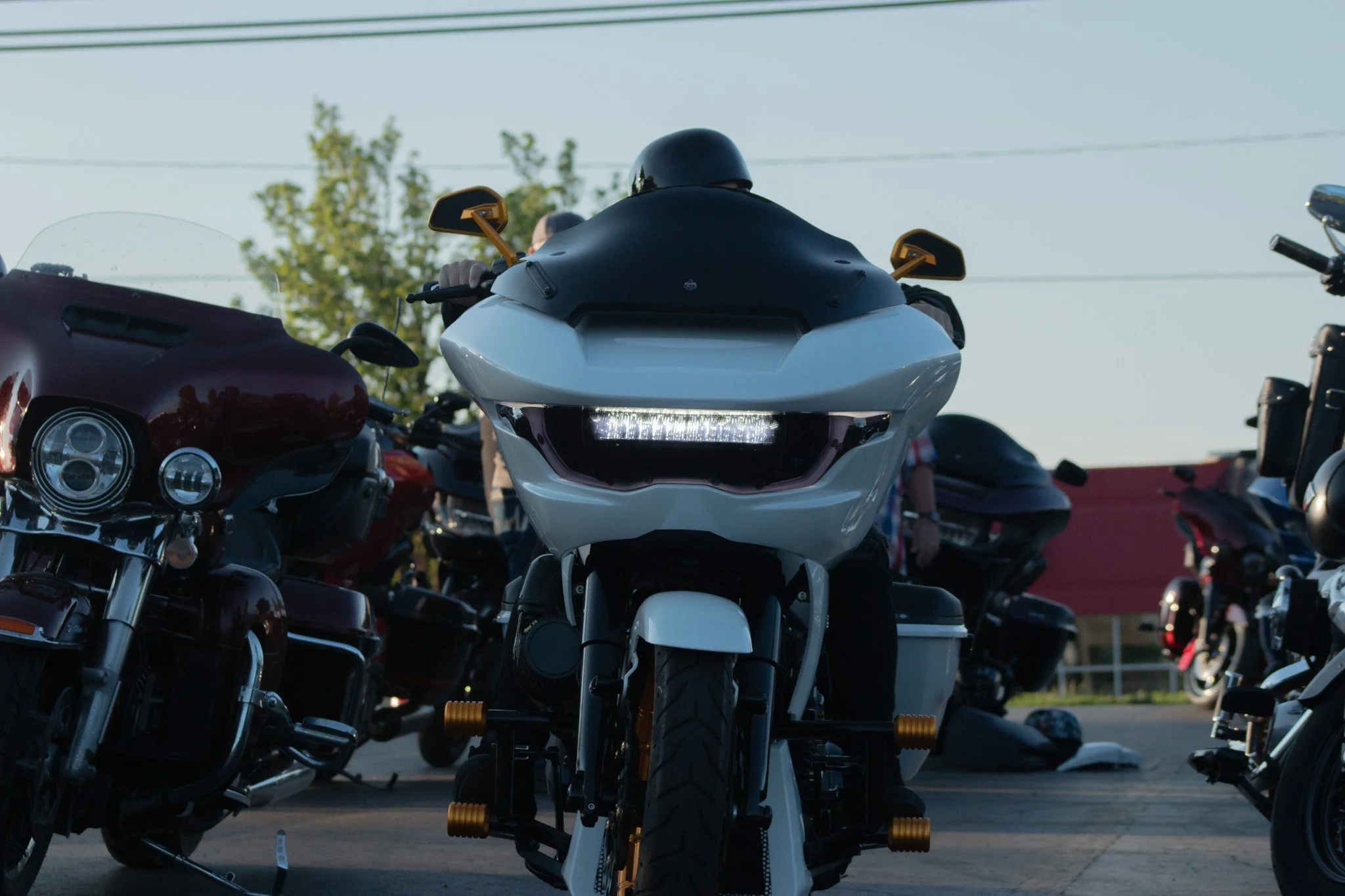 Several motorcycles parked closely together outdoors, with a focus on a white motorbike in the center, surrounded by other bikes including a dark red and a black motorcycle, under a clear sky with trees in the background.