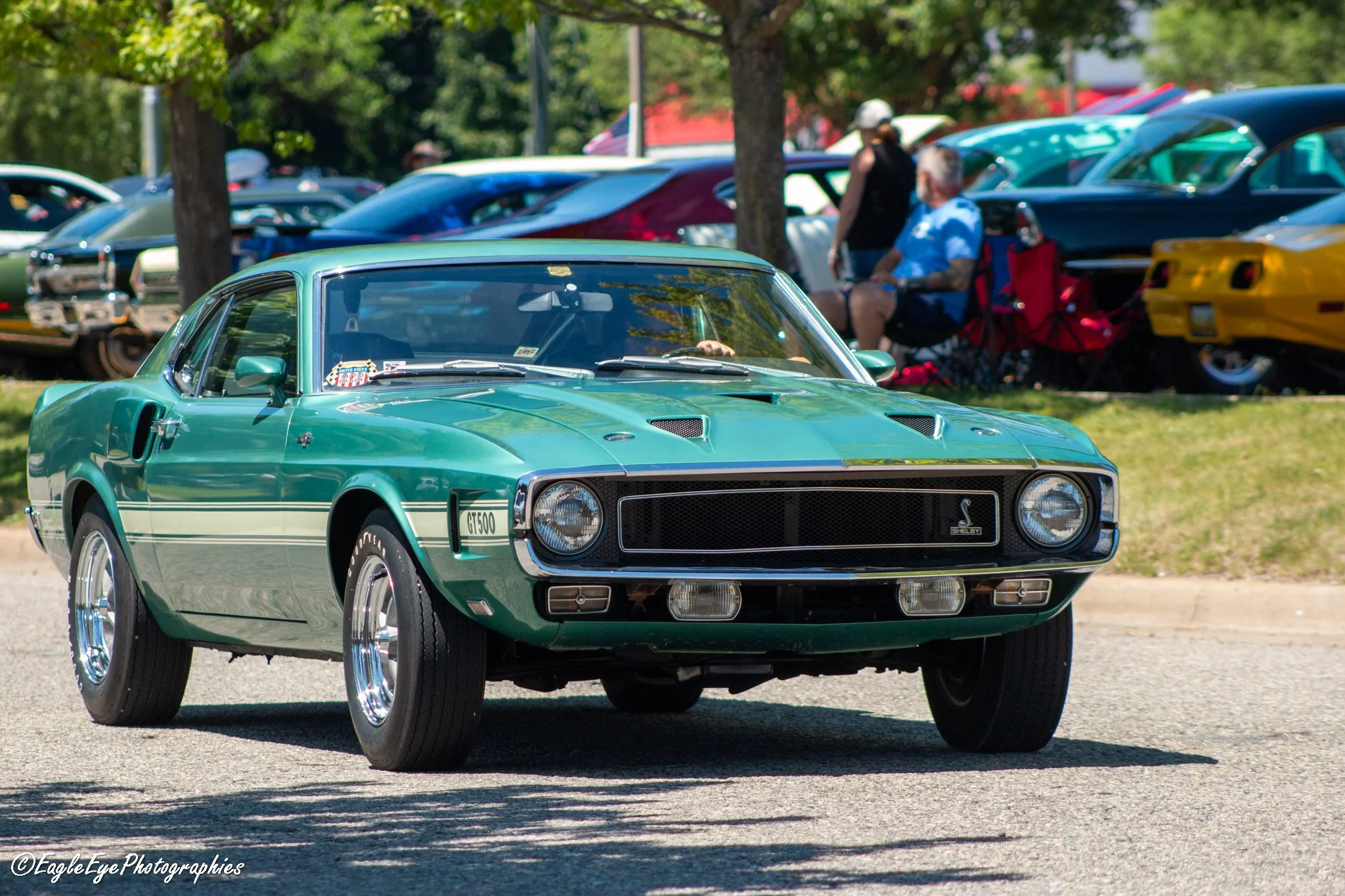 A vintage green Shelby GT500 muscle car parked on a grass area at a car show, with people and other classic cars in the background.